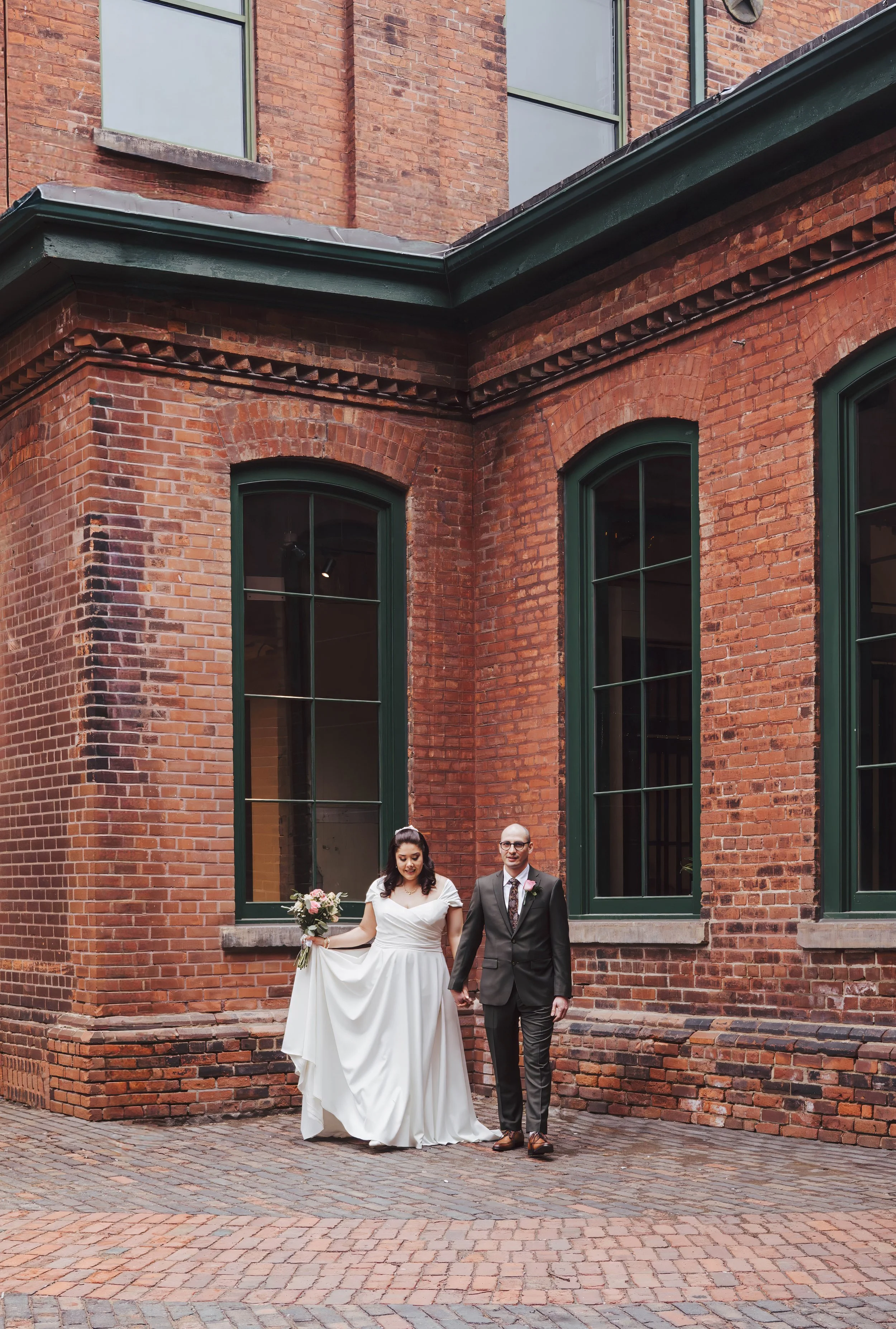 A bride and groom walking hand in hand outside on a cobblestone street in front of a red brick building with large green framed windows.