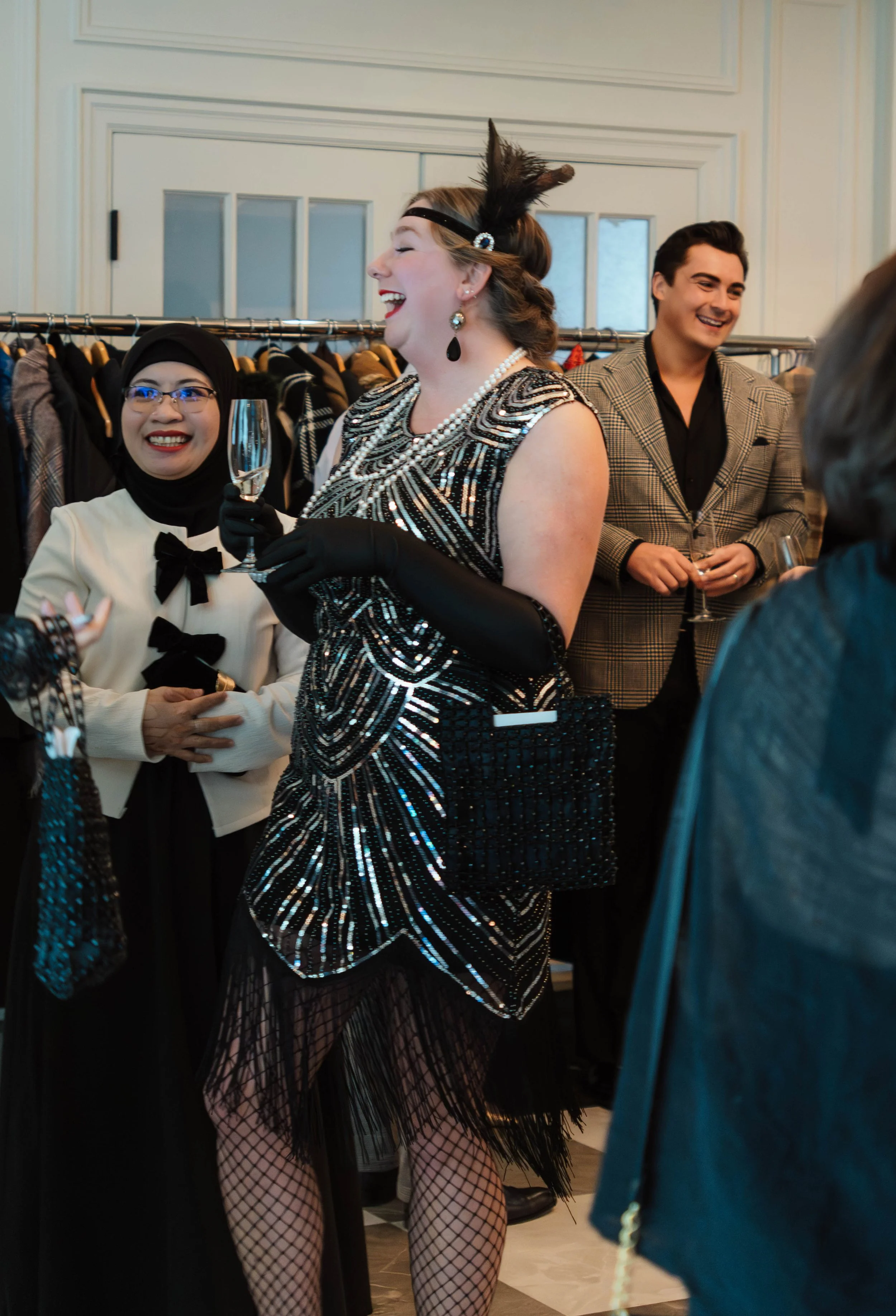 Group of people dressed in vintage 1920s attire at a social gathering, with a woman in the center wearing a black and silver beaded flapper dress, black gloves, and a headband with a feather, holding a champagne glass, smiling and talking to others.