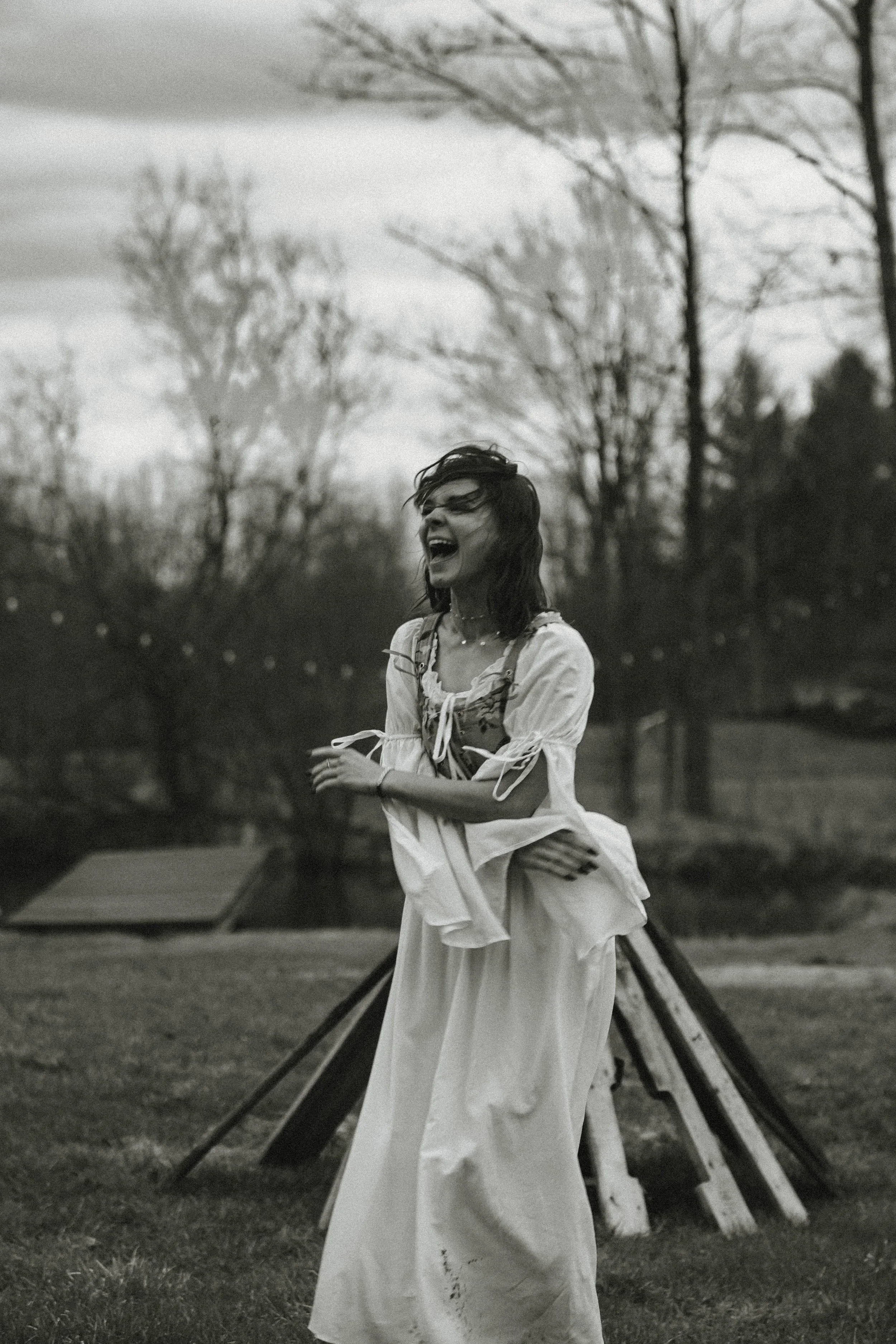 A woman laughing and standing outdoors on a grassy area, with leafless trees and a wooden structure in the background, in black and white.