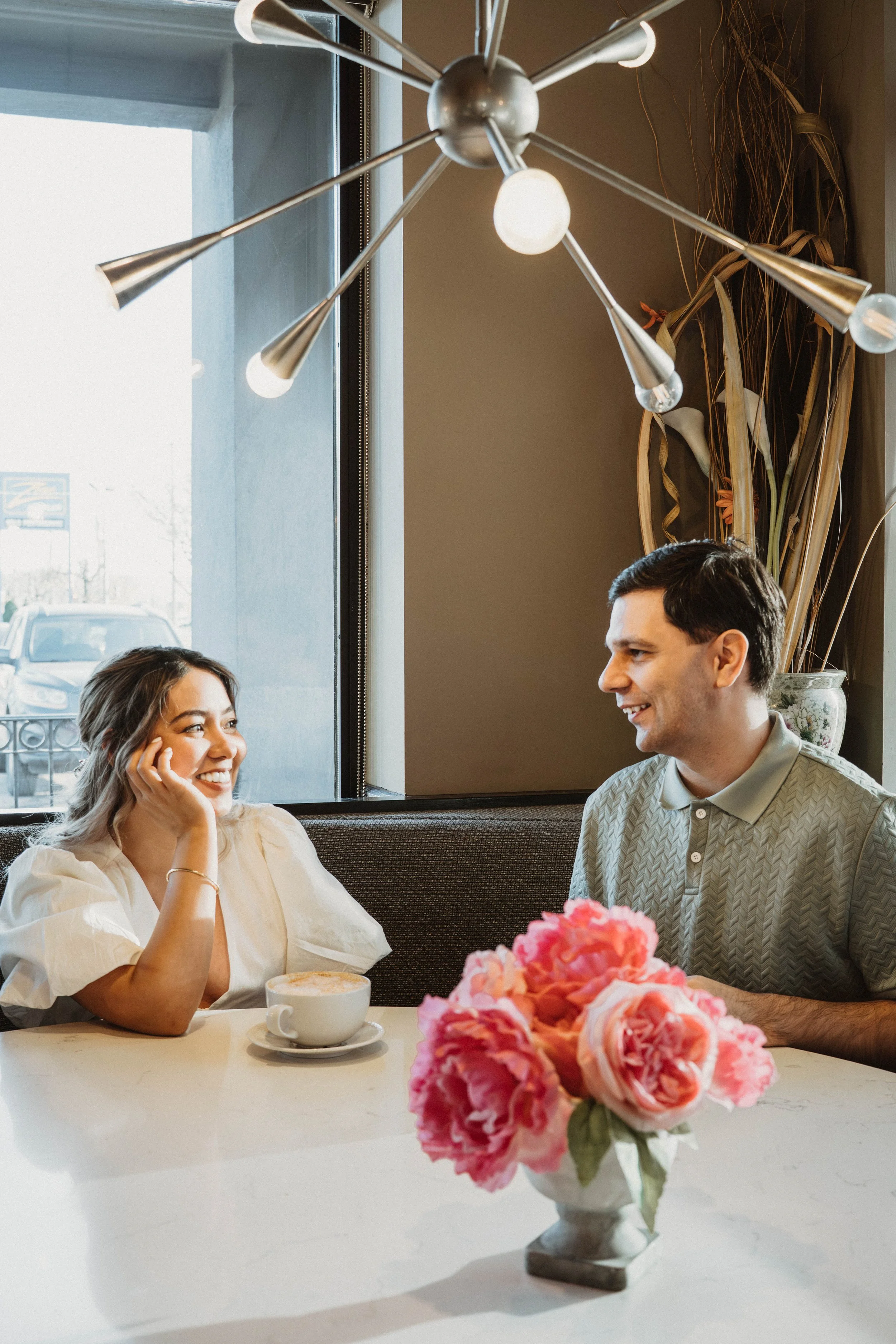 A woman and a man sitting at a restaurant table having a conversation. The woman has light brown hair, is wearing a white blouse, and is smiling while touching her face. The man has dark hair, is wearing a light-colored, patterned shirt, and is smili