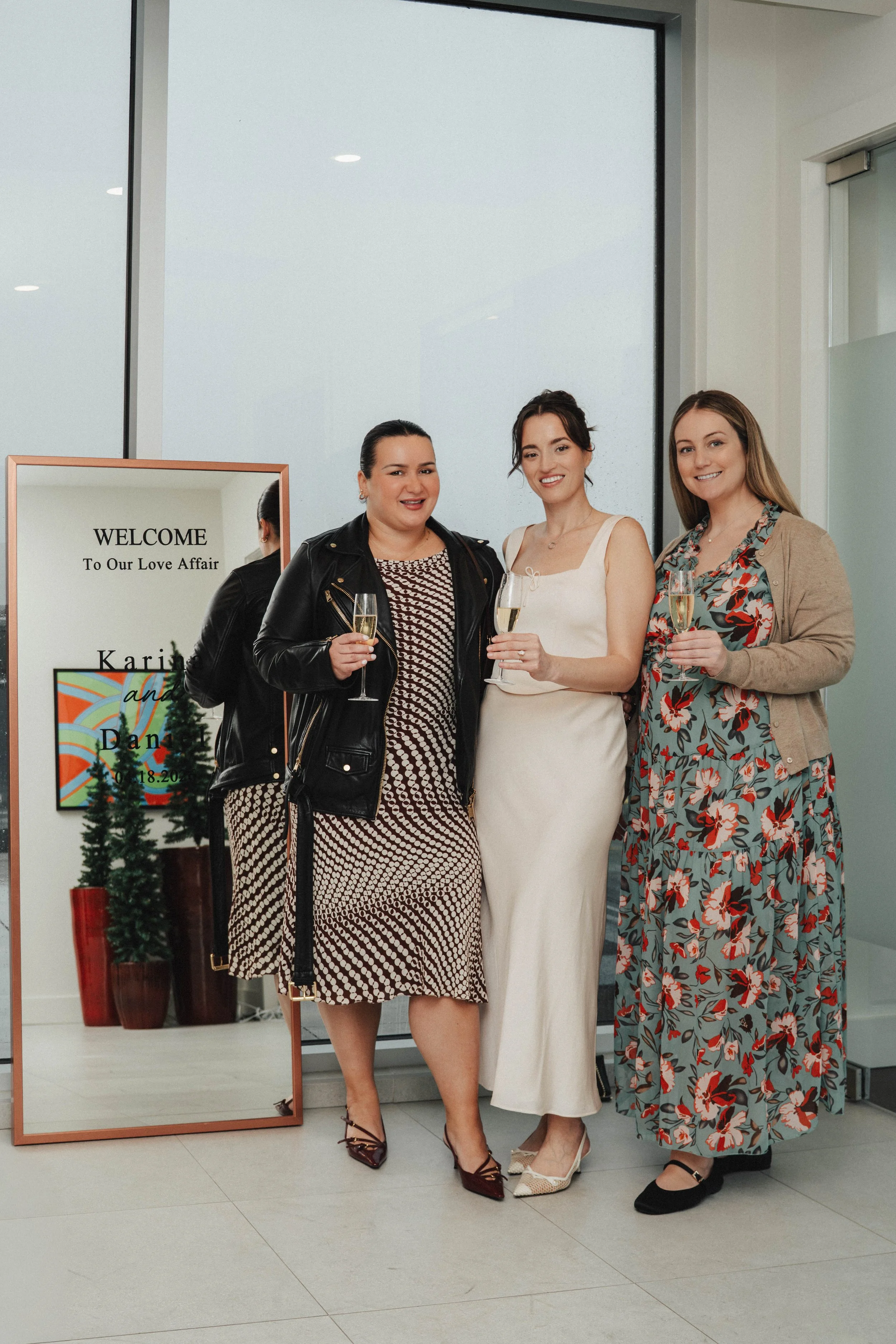 Three women dressed in formal attire, holding glasses of champagne, standing in front of a mirror and wall with a welcome sign for a wedding called 'Our Love Affair'.