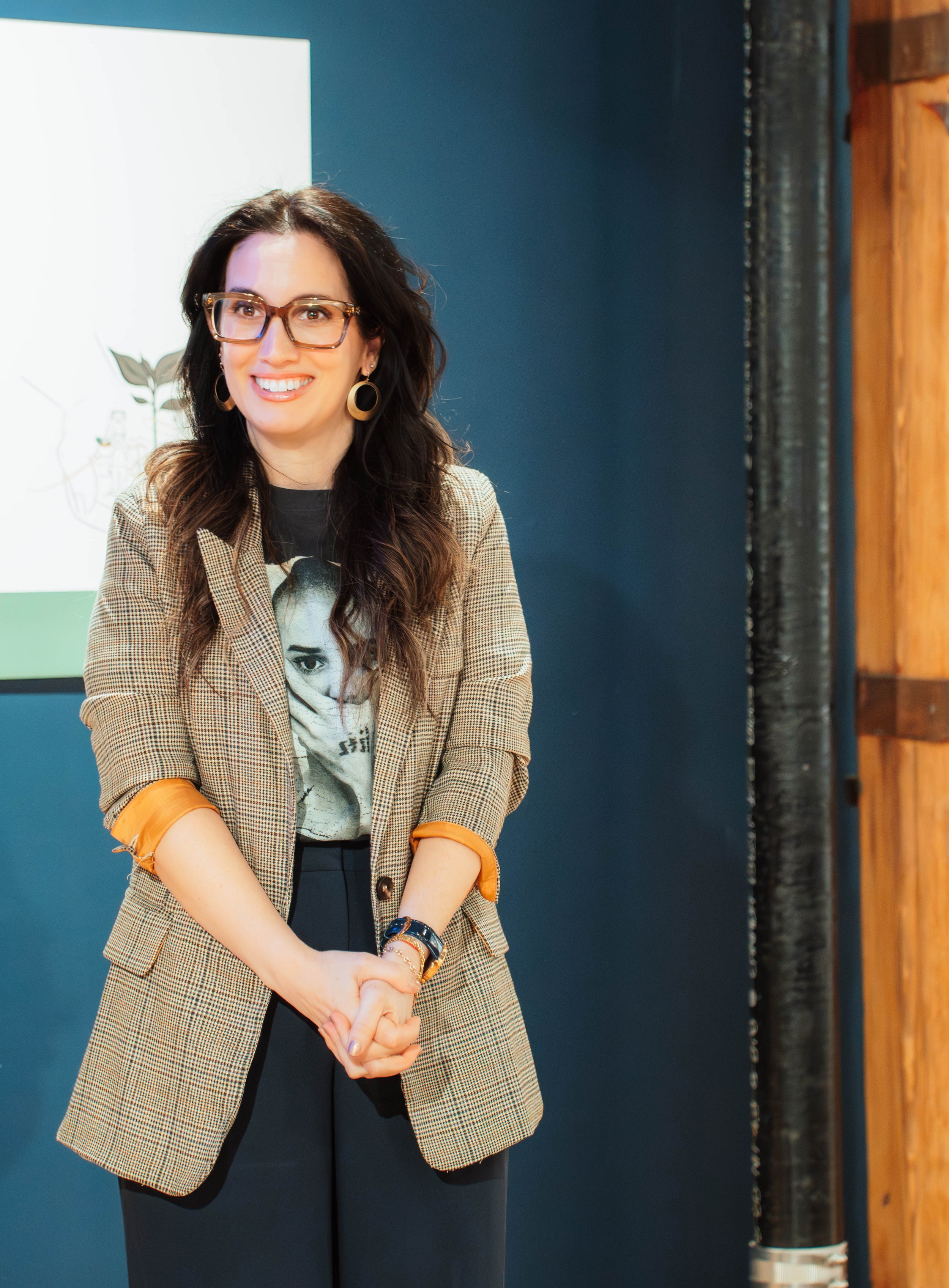 A woman with long dark hair, glasses, and earrings, smiling and standing in front of a dark blue wall with a presentation screen behind her.