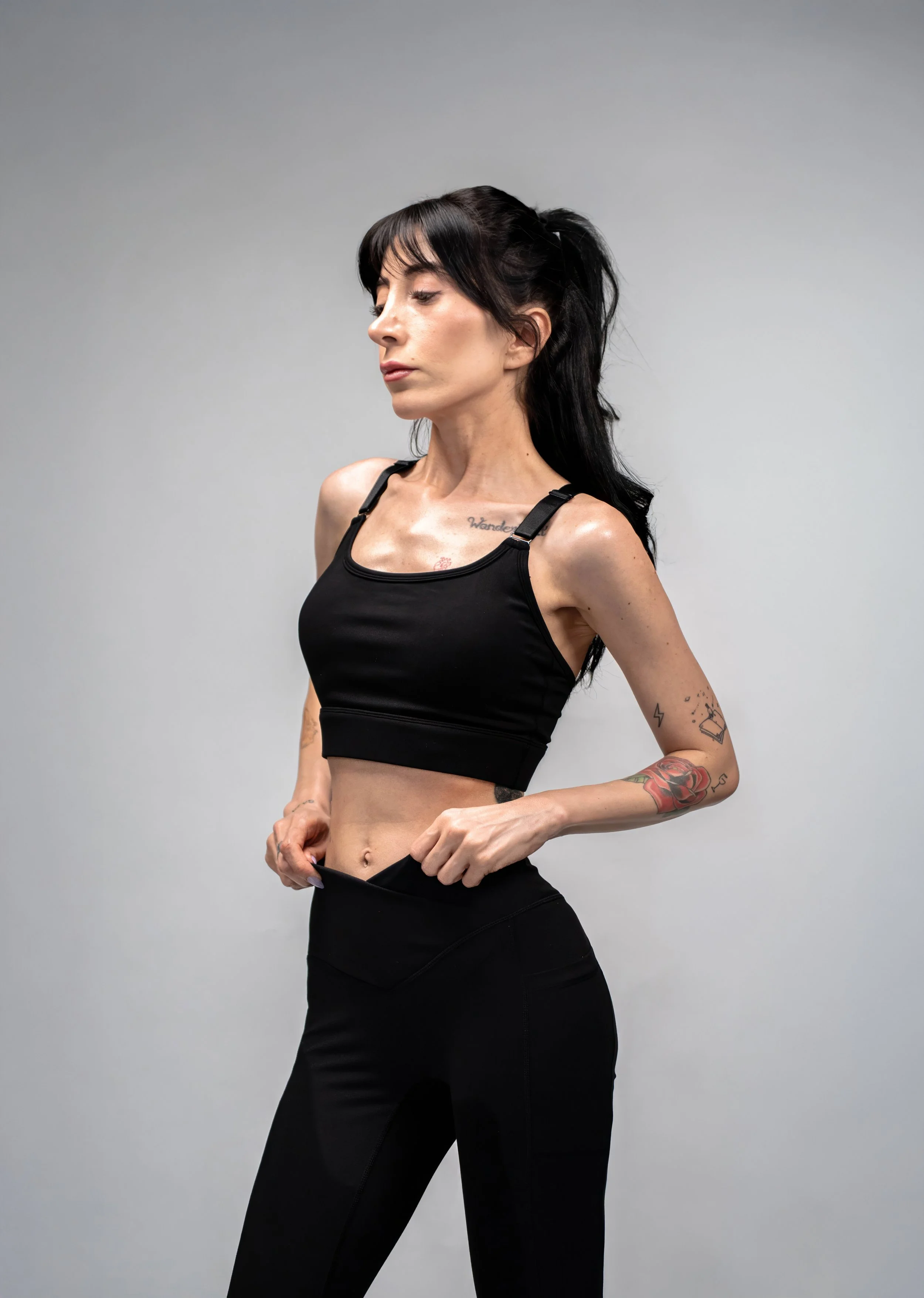 A woman with black hair in a high ponytail, wearing a black sports bra and black workout pants, standing against a plain gray background.