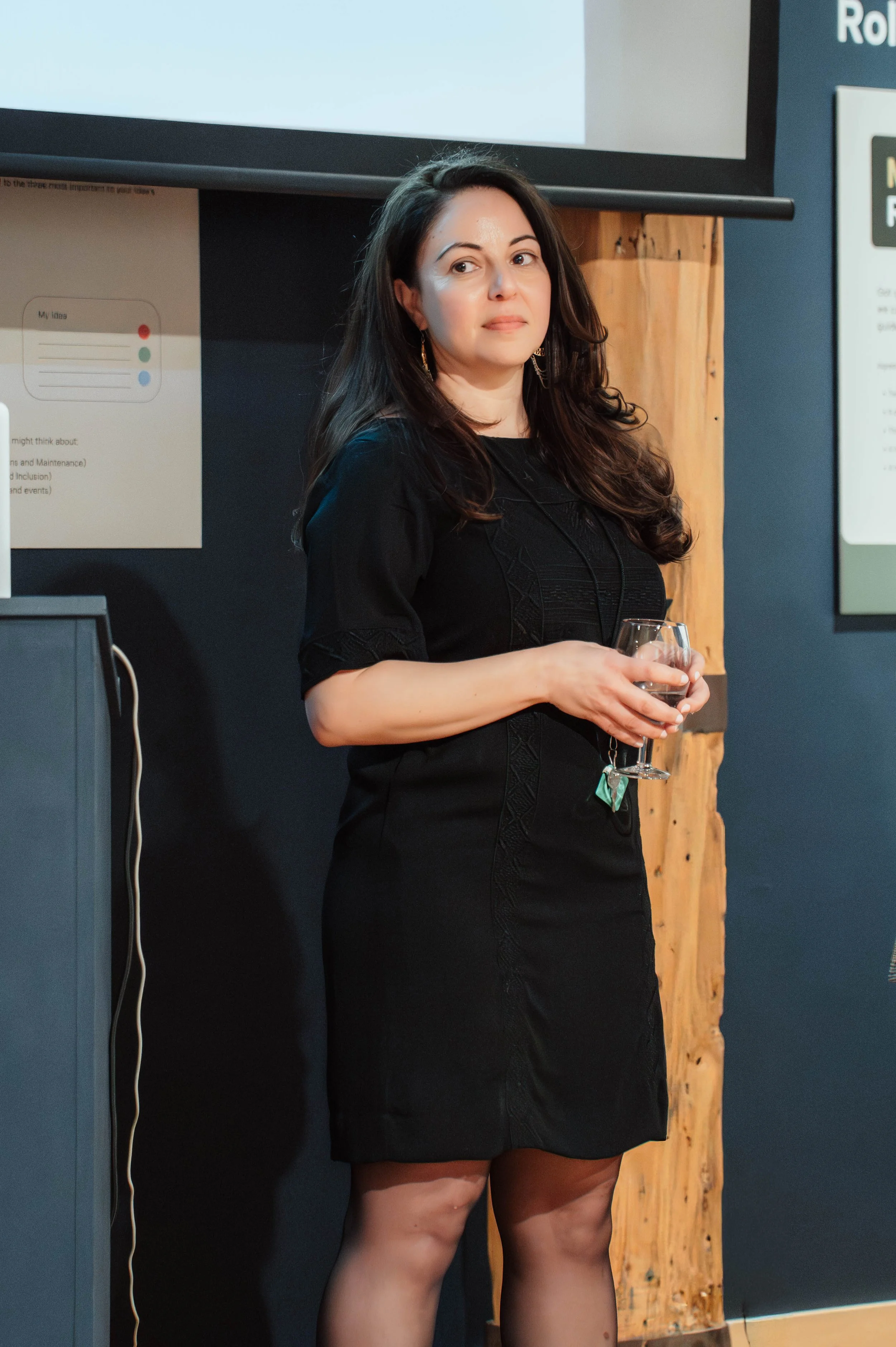 A woman with long dark hair wearing a black dress and earrings, holding a glass of wine in a room with dark walls, a wooden panel, and presentation screens.