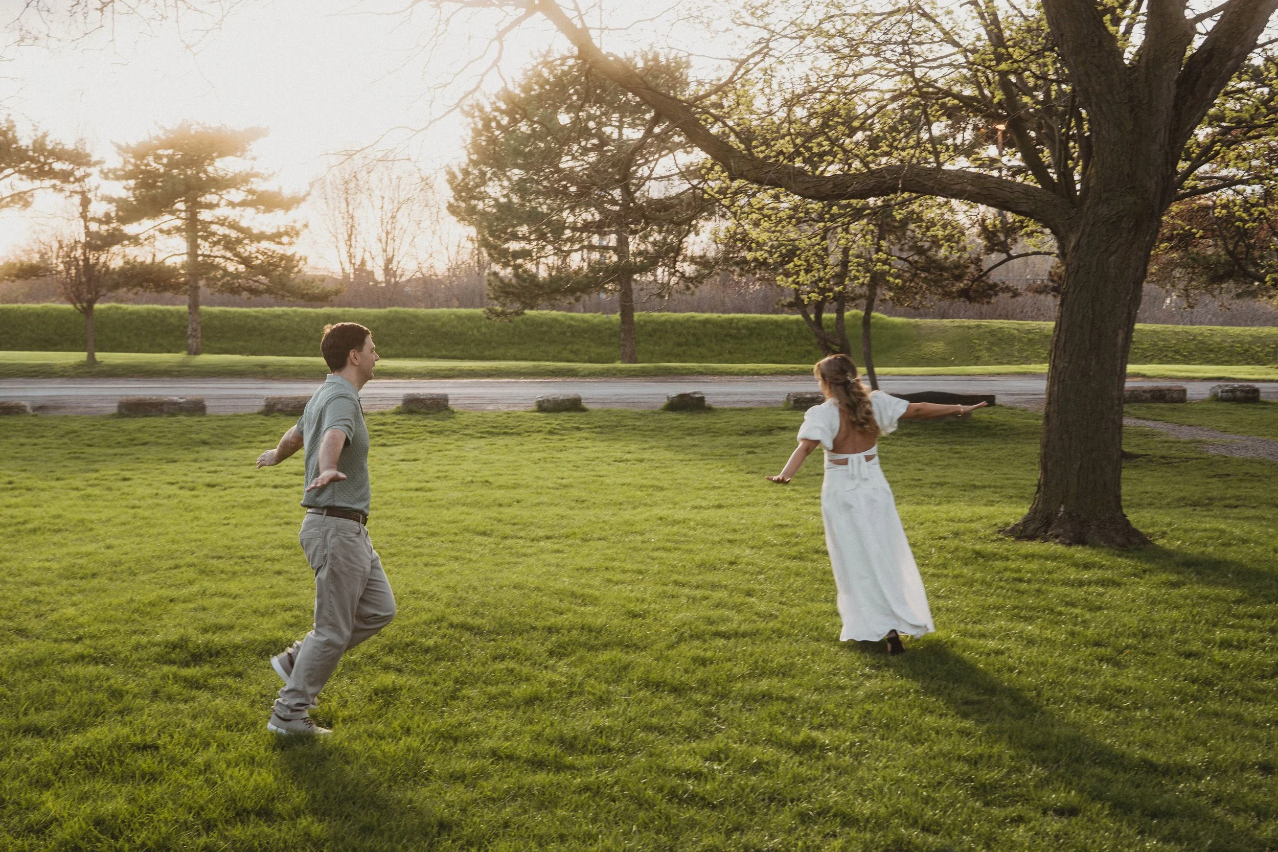 A man and a woman are playing or dancing outdoors in a park at sunset, with trees and a grassy area in the background.
