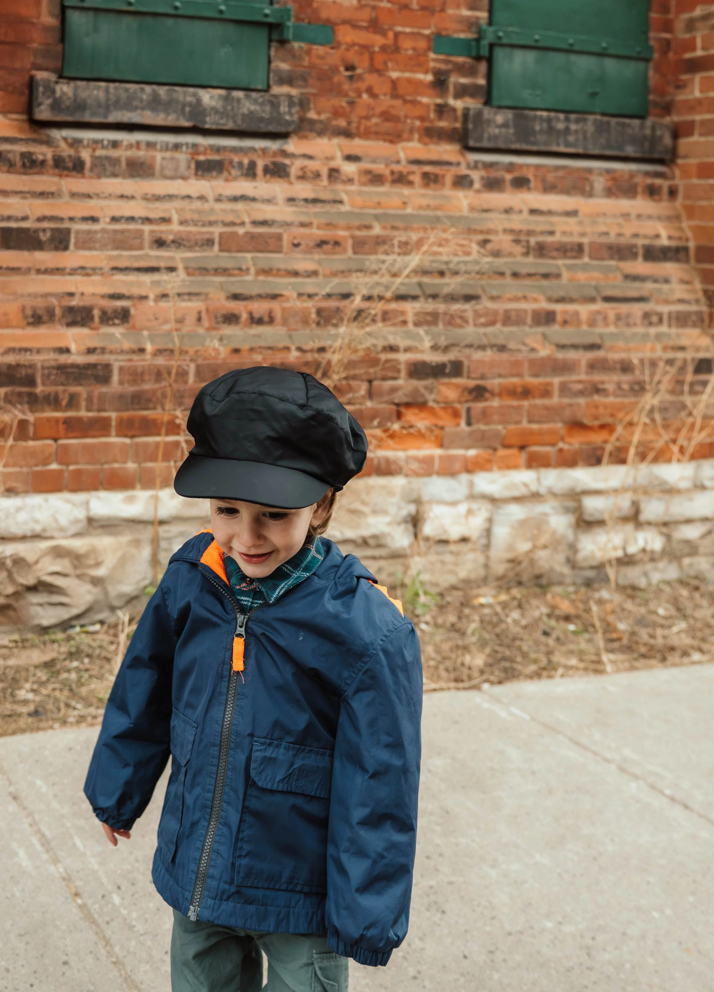 A young boy wearing a black cap, blue jacket with orange accents, and green pants walking outdoors on a sidewalk in front of a brick wall with green shutters.