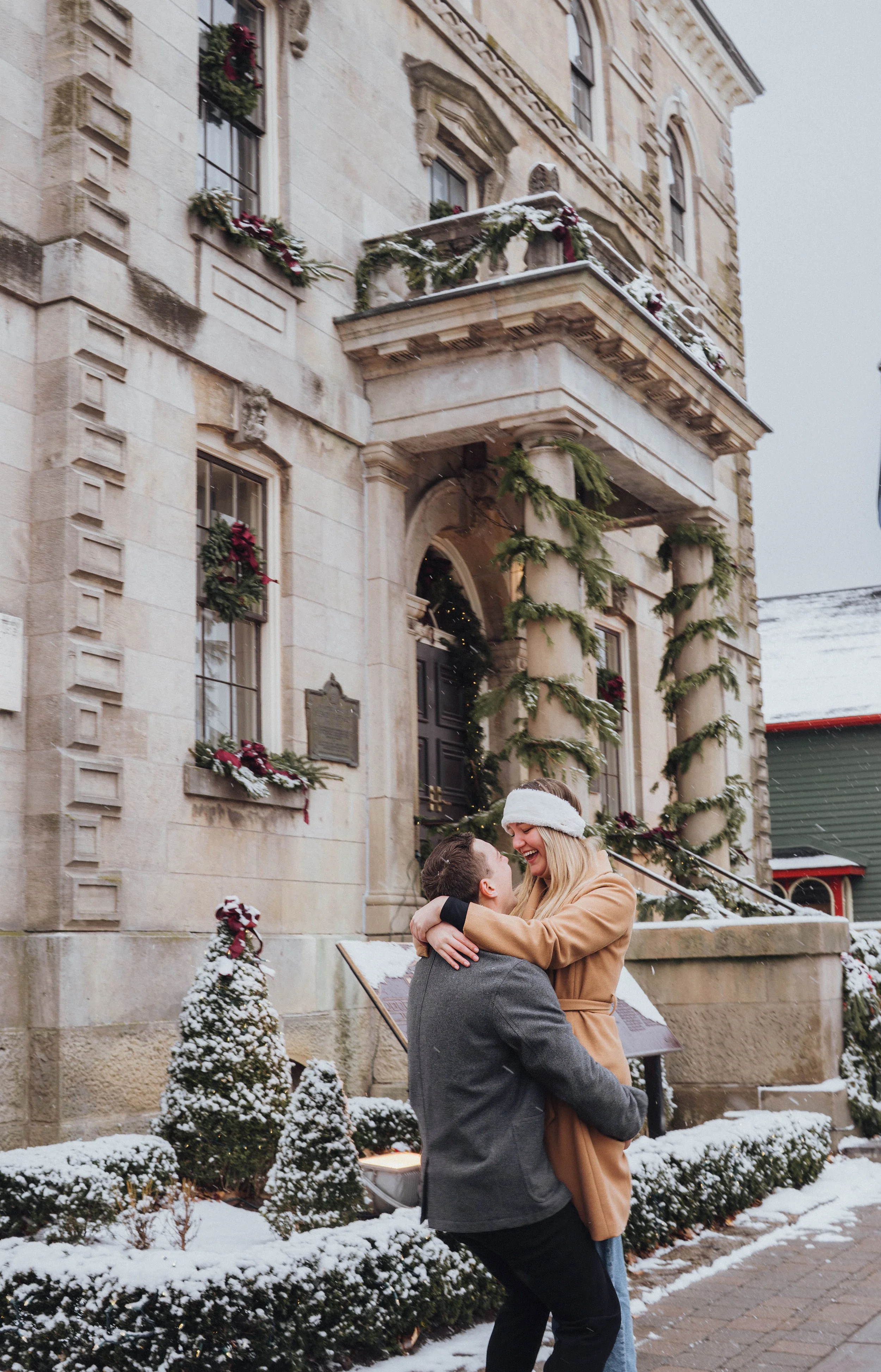 Christmas couple photography in Niagara-on-the-Lake featuring a playful candid moment in the snow surrounded by festive holiday greenery and Christmas decorations.