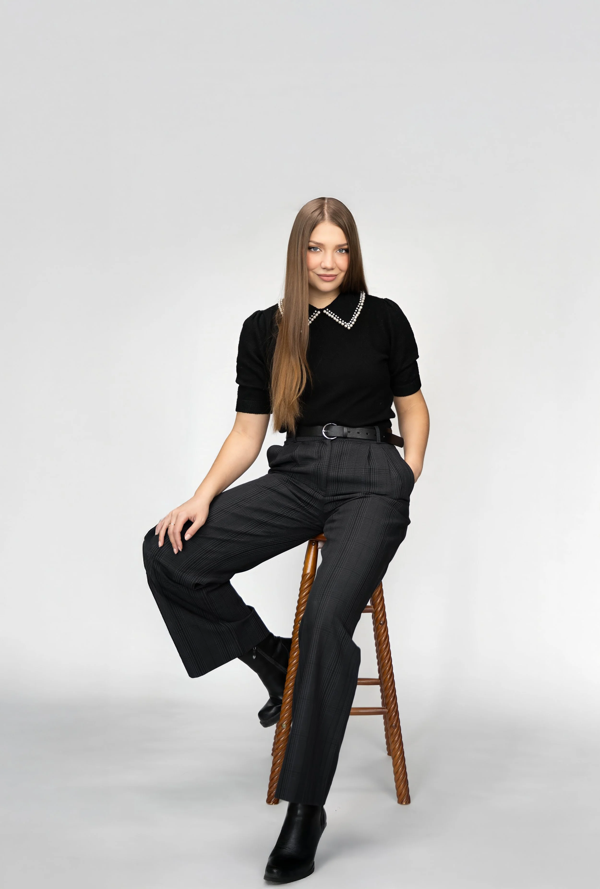 A young woman with long brown hair wearing a black blouse with pearl collar details, plaid black pants, and black boots, sitting on a wooden stool against a plain white background.