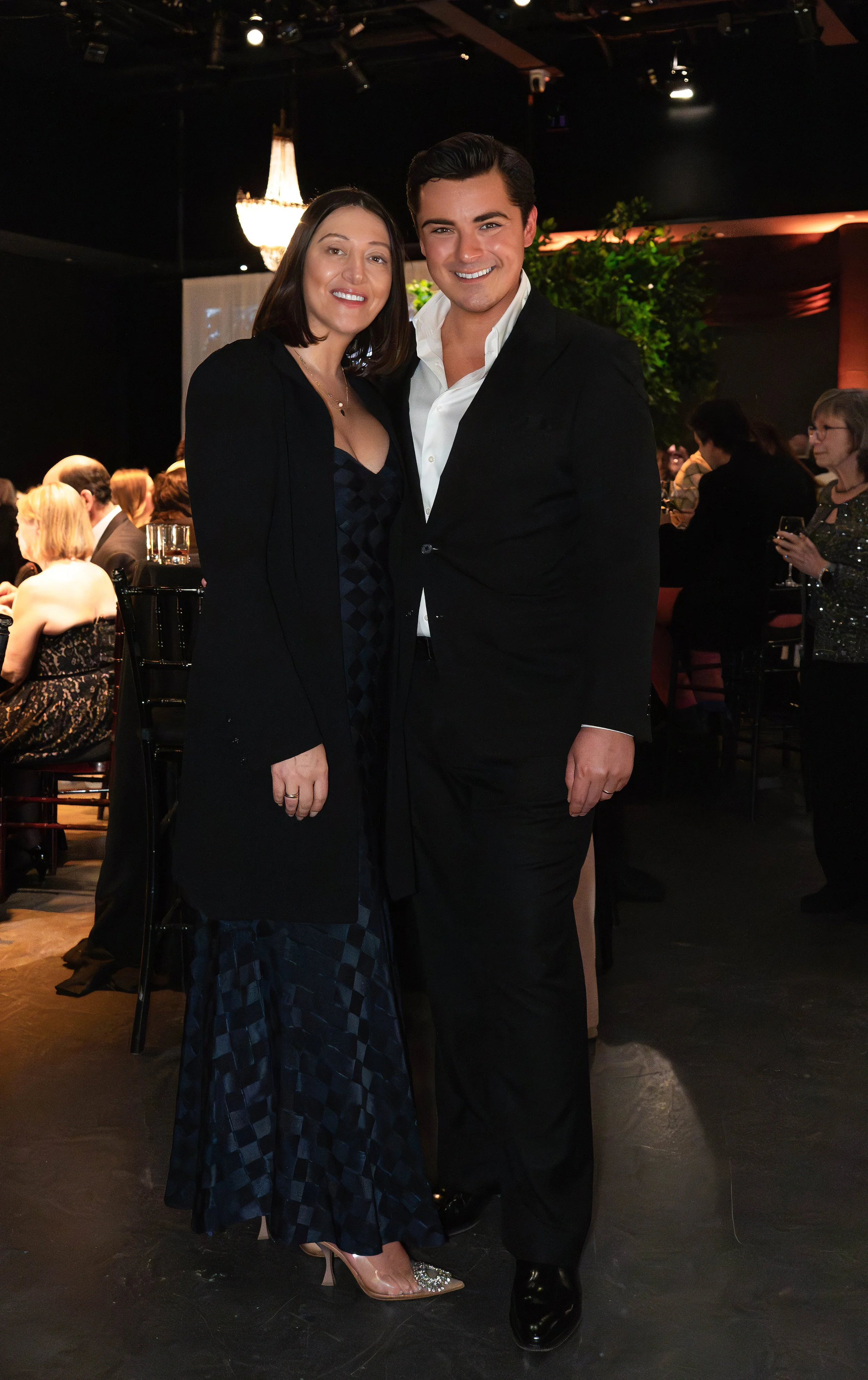 A man and a woman dressed in formal attire standing close together at an indoor event, smiling at the camera, with many people seated at tables in the background.