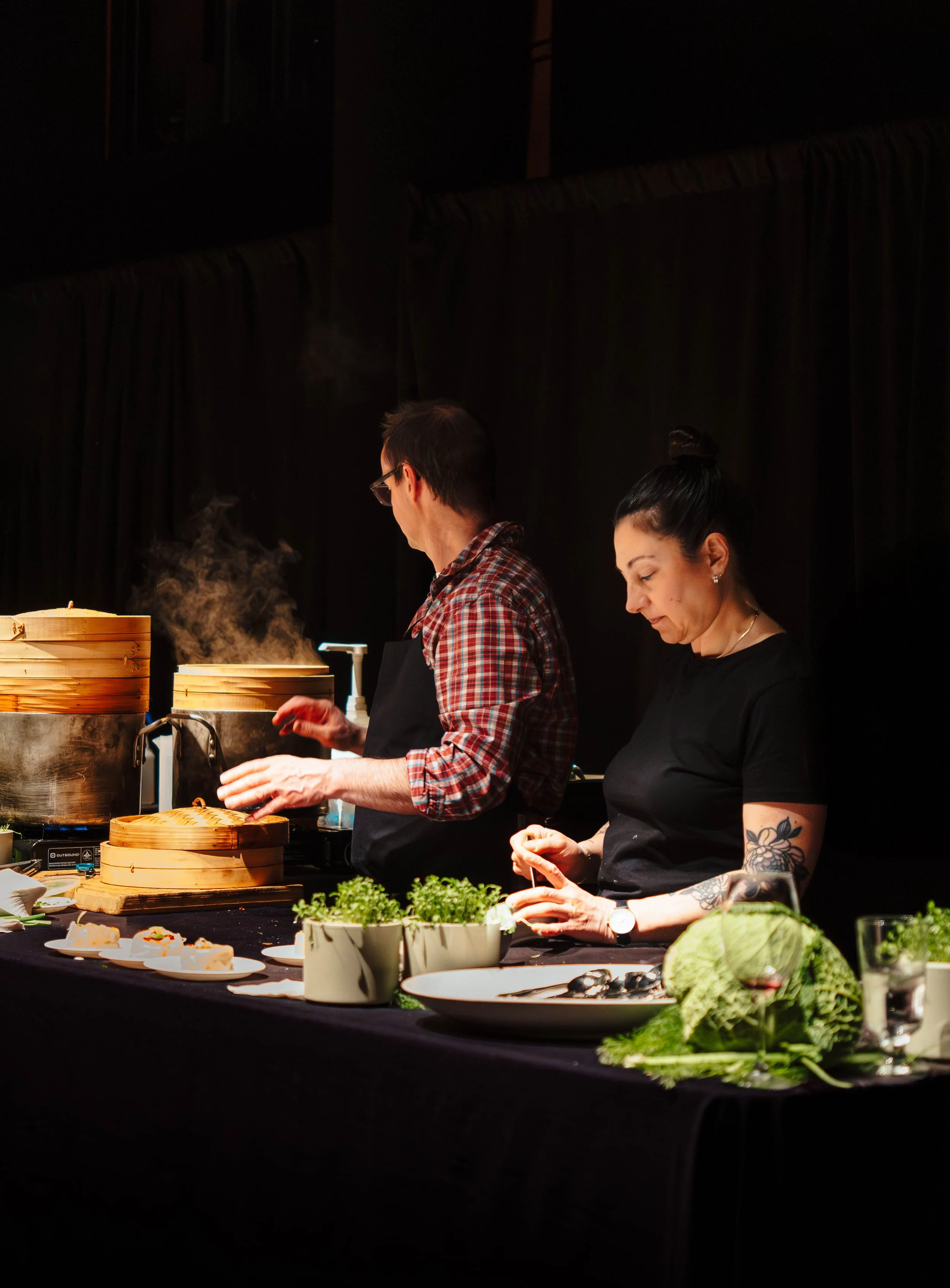 Two chefs, a man and a woman, preparing dishes at a counter with steam rising from bamboo steamers and fresh greens on display.