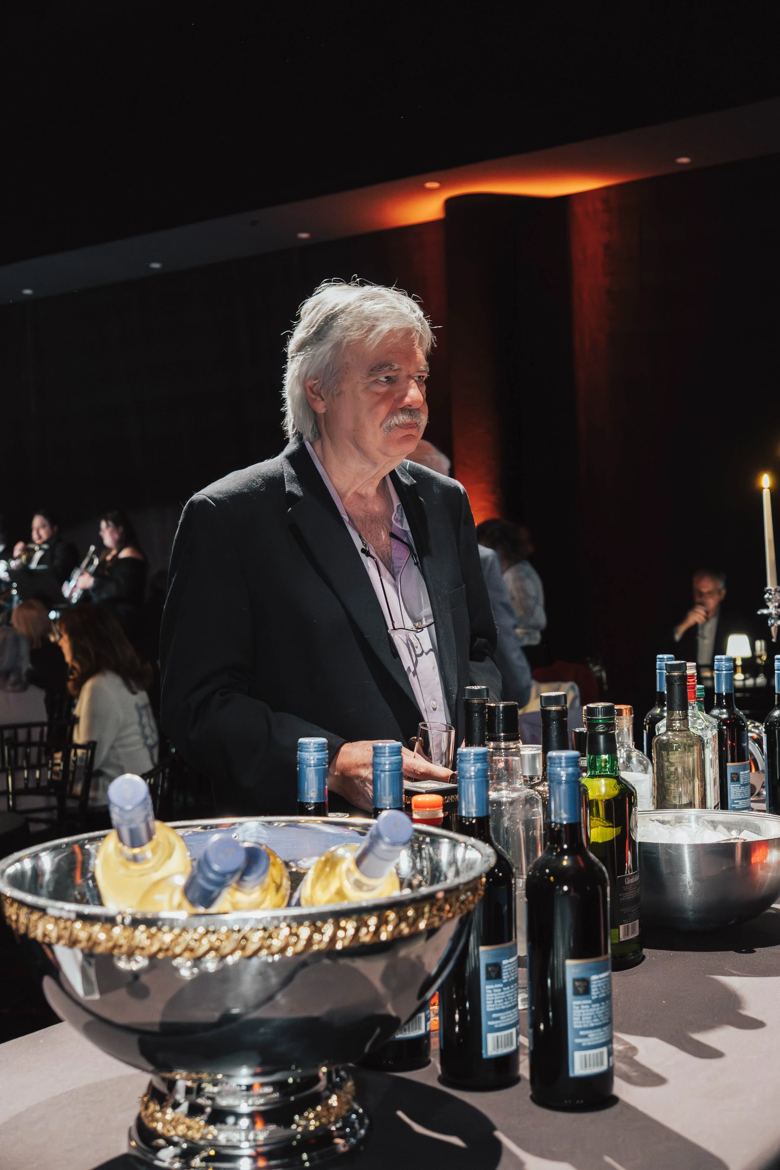 An older man with gray hair and mustache, dressed in a black suit, standing at a bar with various bottles of alcohol in front of him. The setting appears to be a dimly lit event or party.