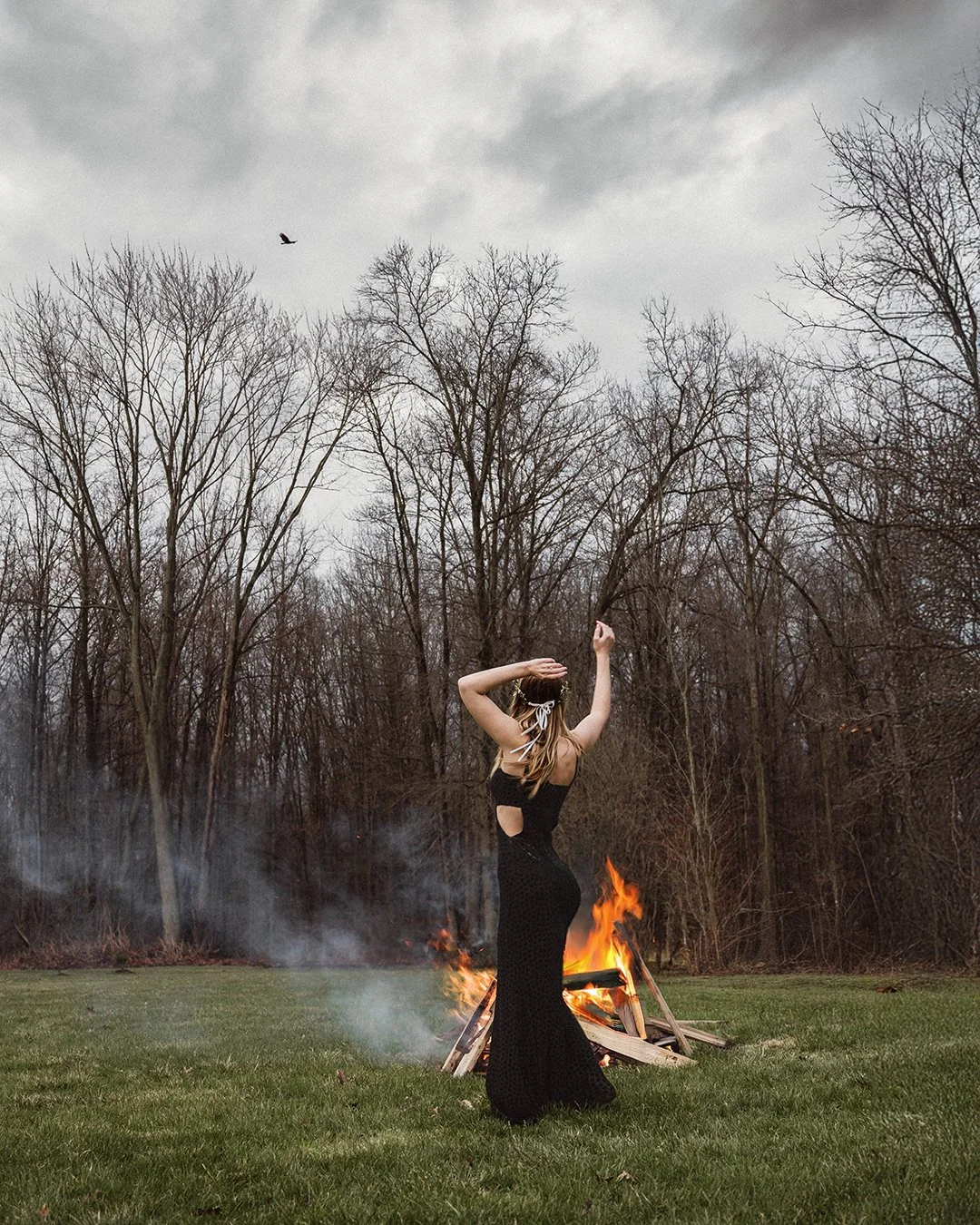 A woman in a black dress with cutouts is dancing outdoors near a bonfire on a grassy field with bare trees and a cloudy sky in the background, with a bird flying overhead.