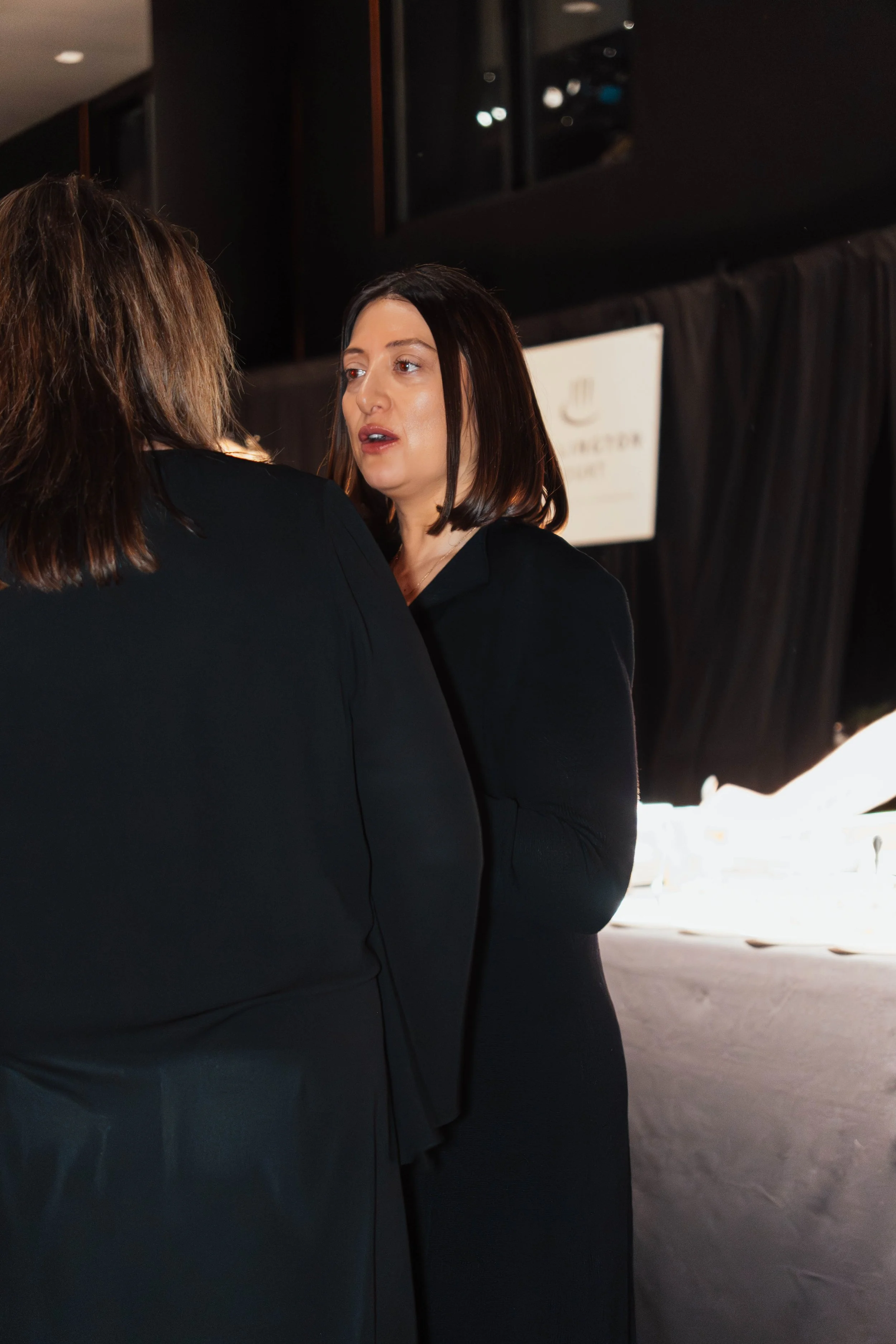 Two women dressed in black engaged in conversation at an indoor event.