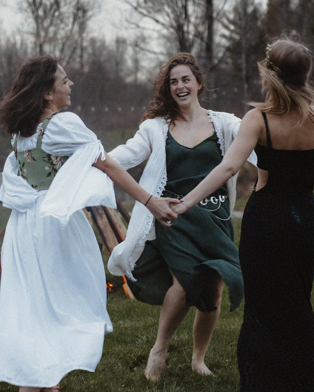 Three women holding hands and dancing outdoors on grass, smiling and enjoying themselves during a gathering or celebration at dusk.