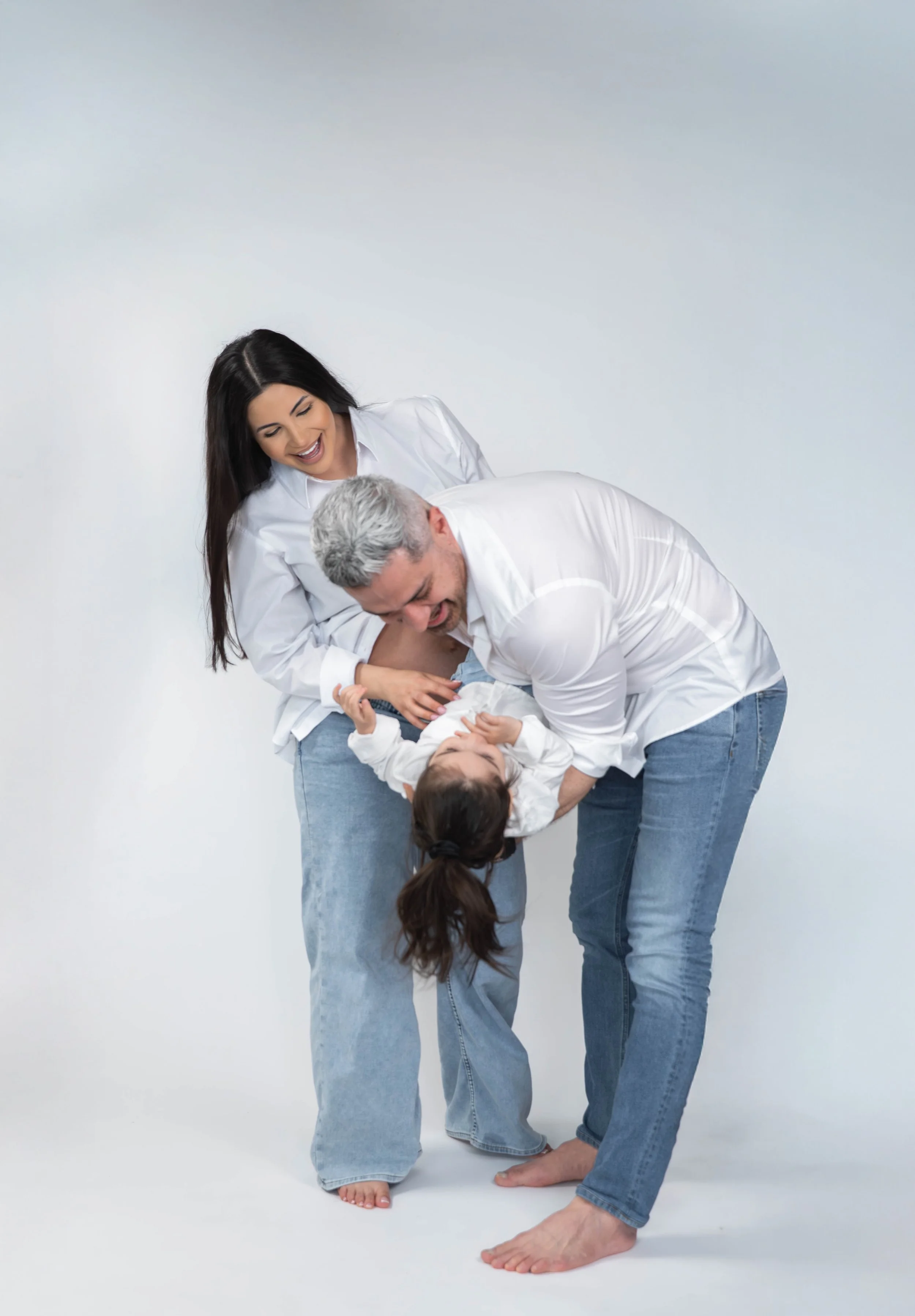 A woman, man, and girl laughing and playing together in a playful, affectionate moment against a plain background, dressed in white shirts and jeans.