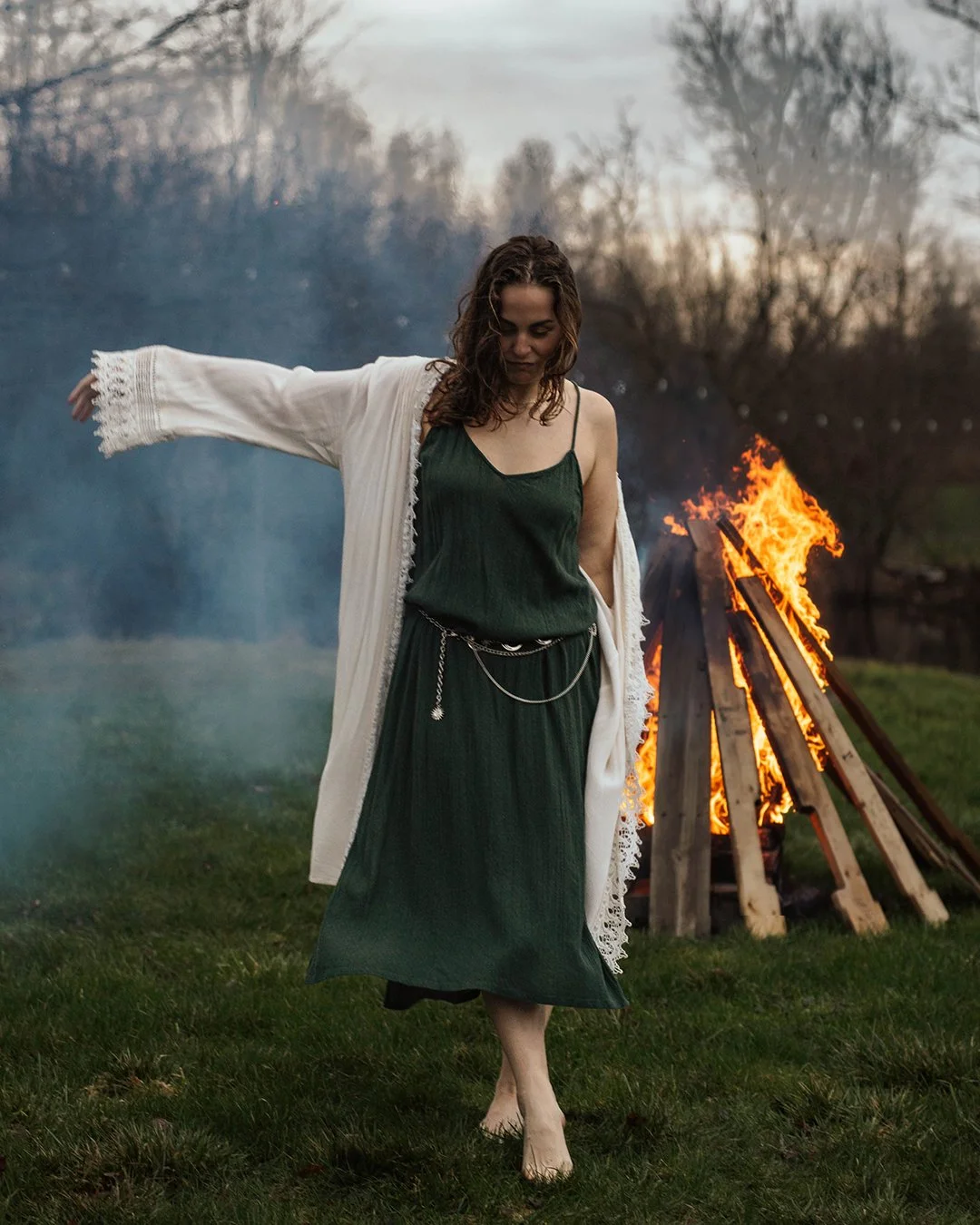 A woman in a dark green dress and white coat with lace trim dancing barefoot near a large bonfire outdoors during dusk.