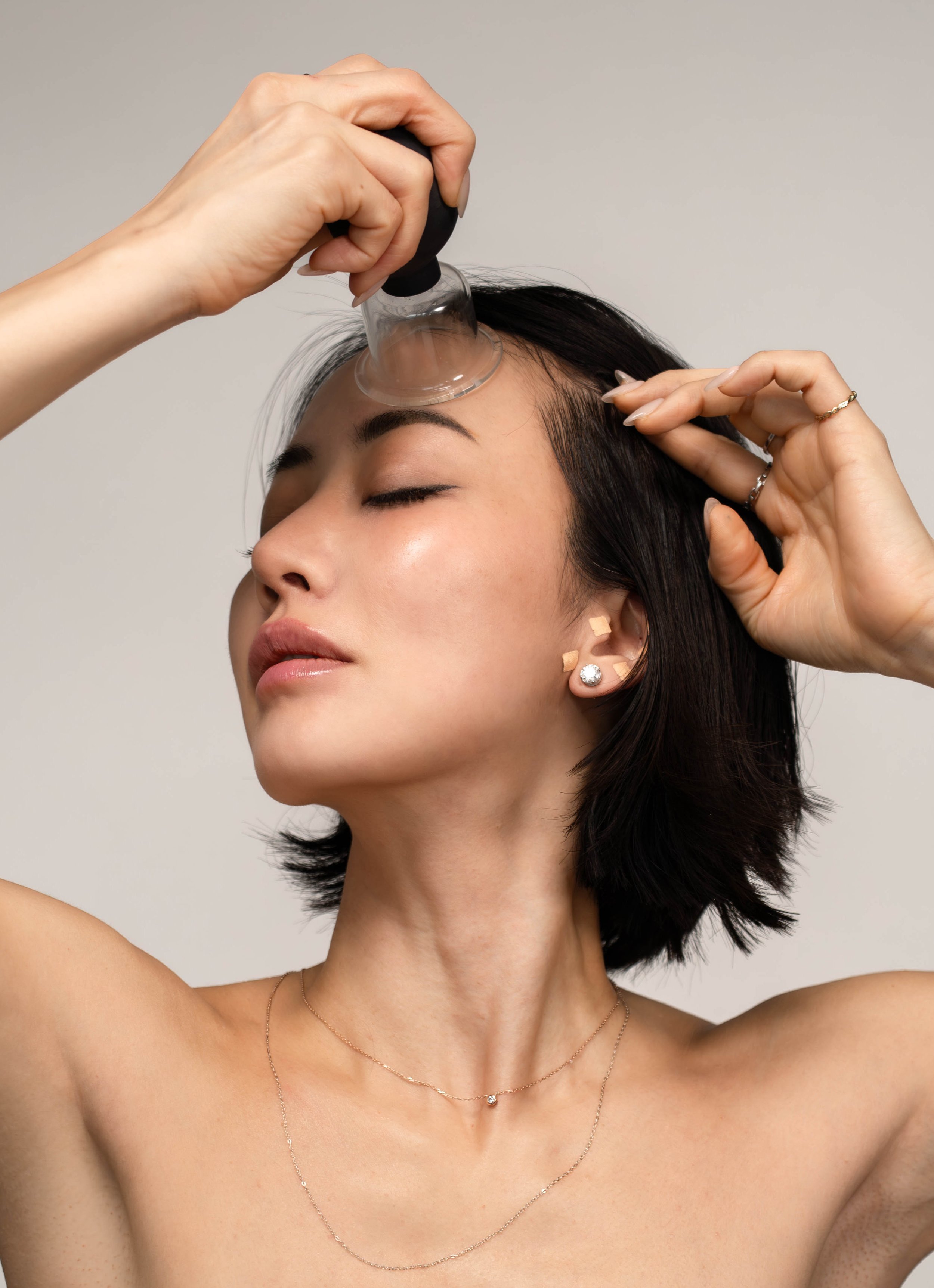 A woman with short black hair and jewelry is undergoing a facial treatment, with her eyes closed. A practitioner is using a glass suction cup device on her forehead, applying gentle pressure.