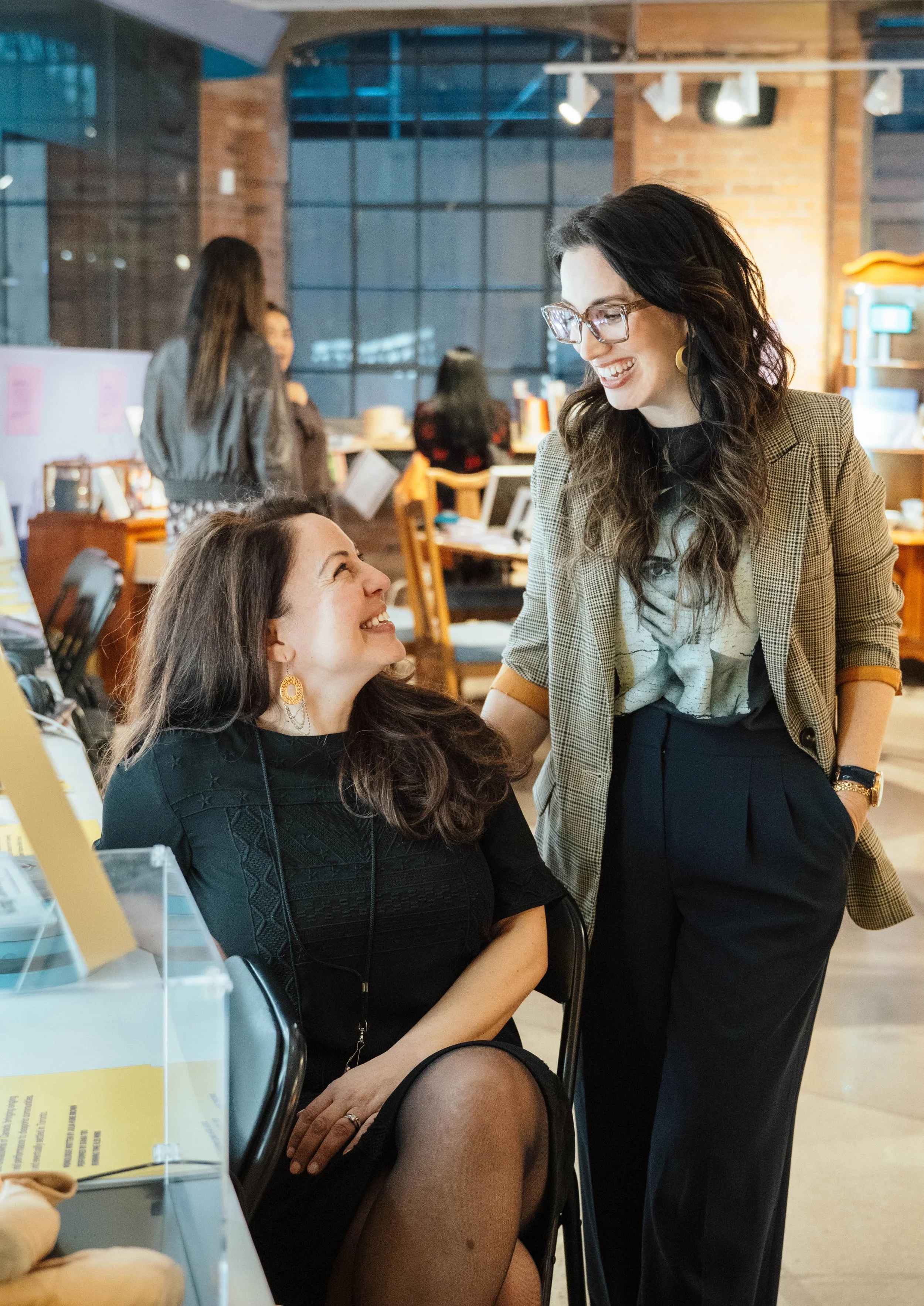 Two women smiling and talking inside a modern, industrial-style cafe or workspace, with others working in the background.