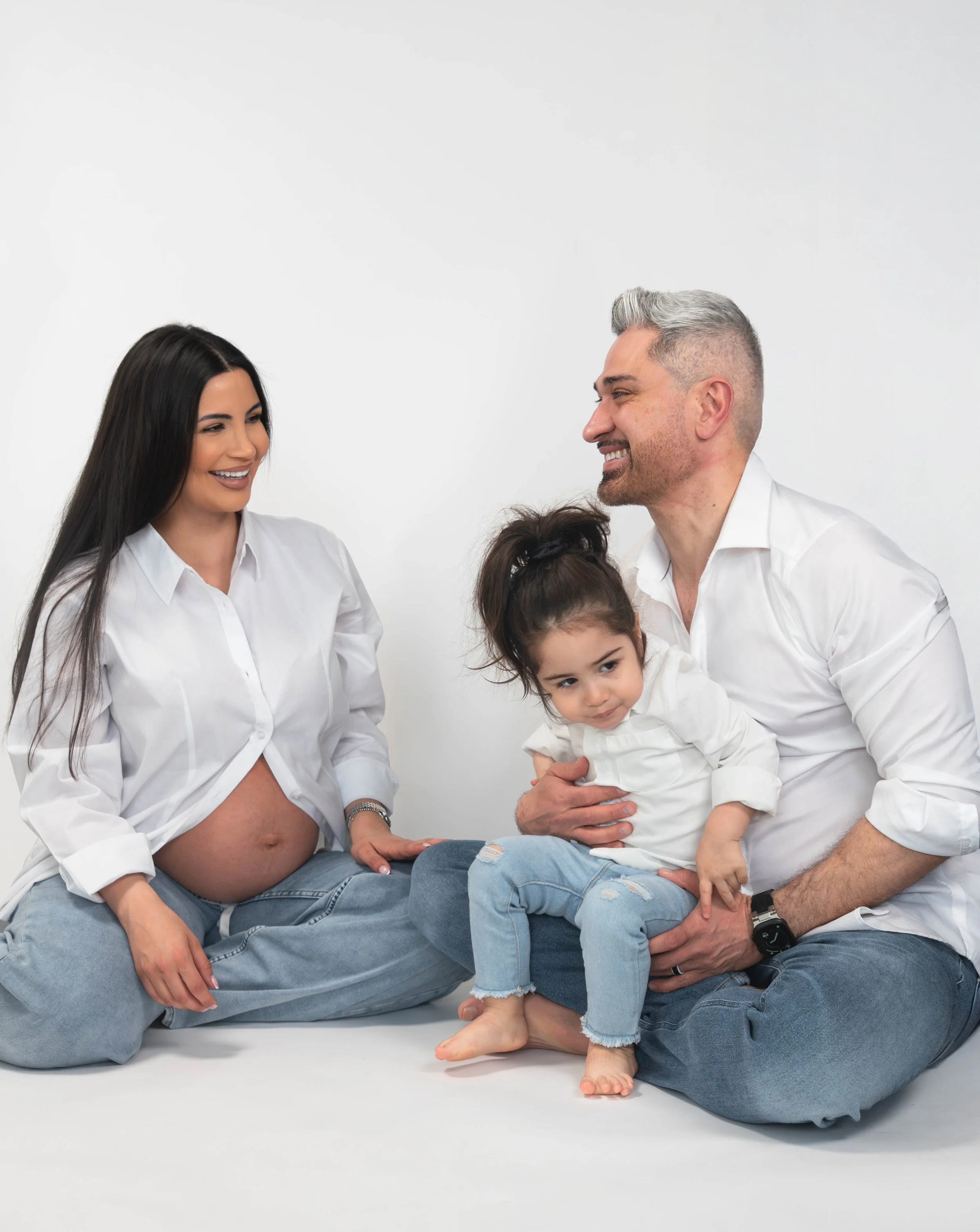 A pregnant woman and a man sitting cross-legged on the floor, smiling and looking at each other, with a young girl sitting on the woman's lap. All are dressed in white shirts and jeans, against a plain white background.