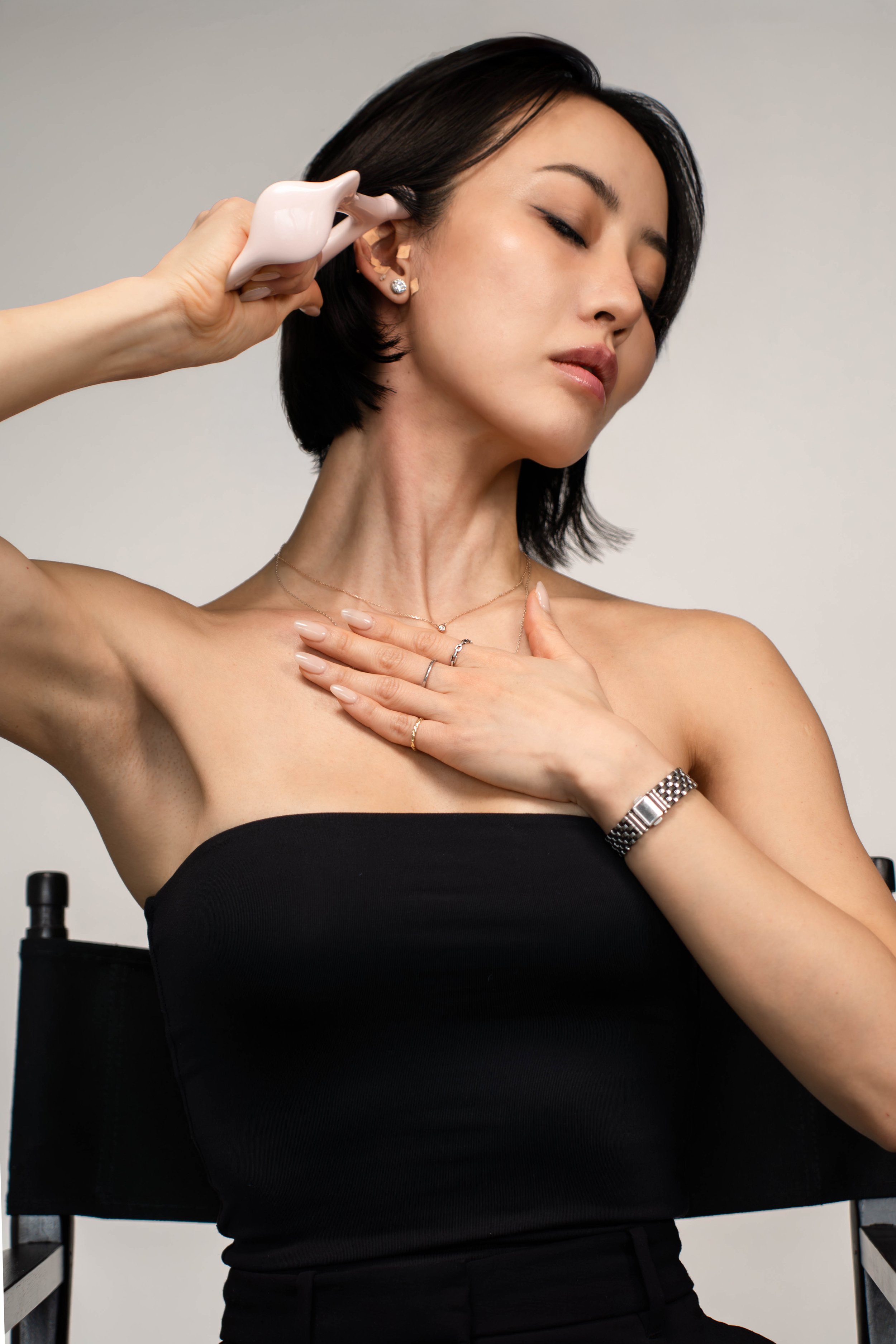 A woman with dark hair in a black strapless top sitting on a director's chair, leaning her head back, with her eyes closed. She is holding a pink ceramic mug to her ear as if listening, with her right hand resting on her chest, adorned with rings and