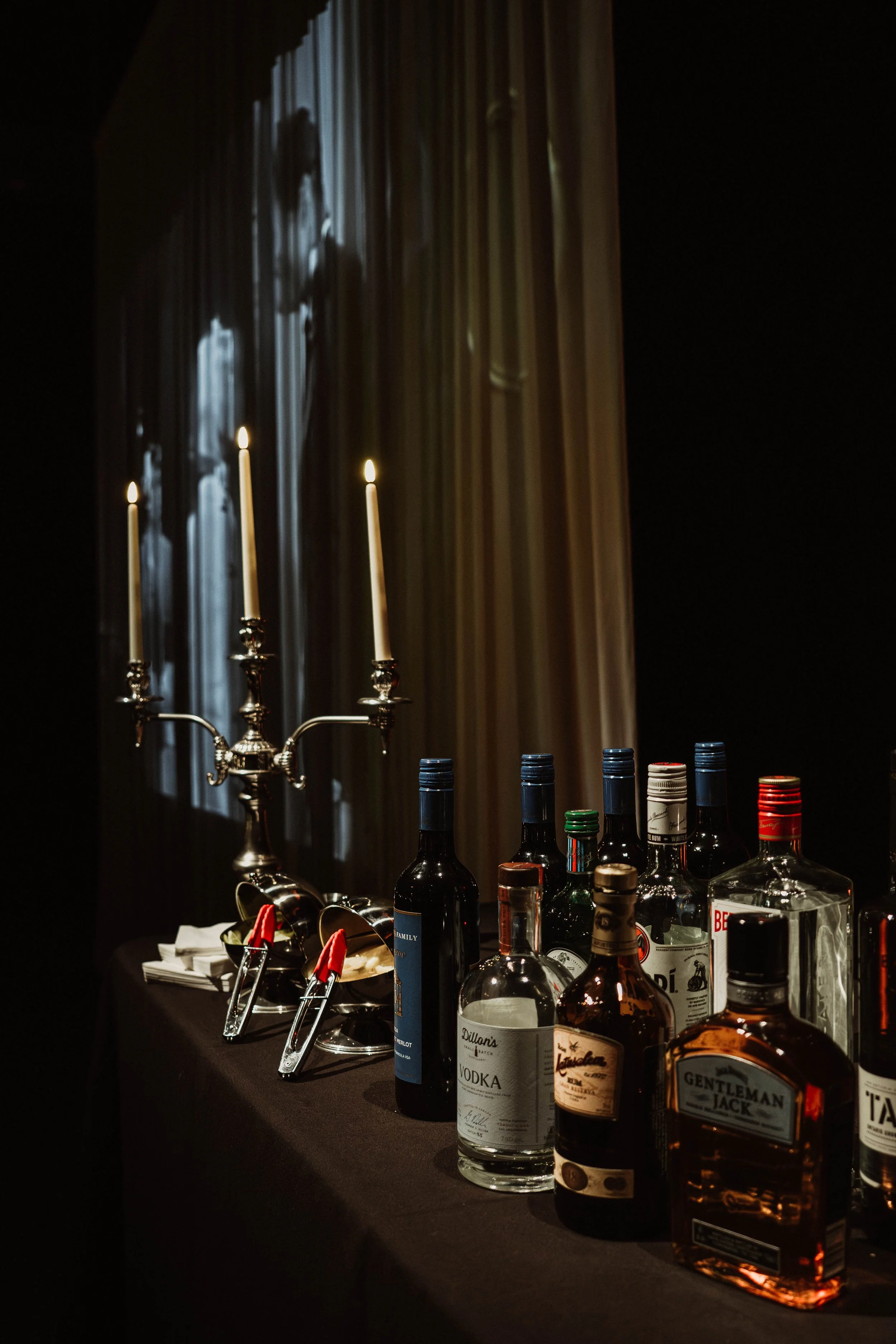 A dimly lit bar table with bottles of alcohol, a silver candle holder with three lit candles, and two clip tongs, with a dramatic shadowy curtain in the background.