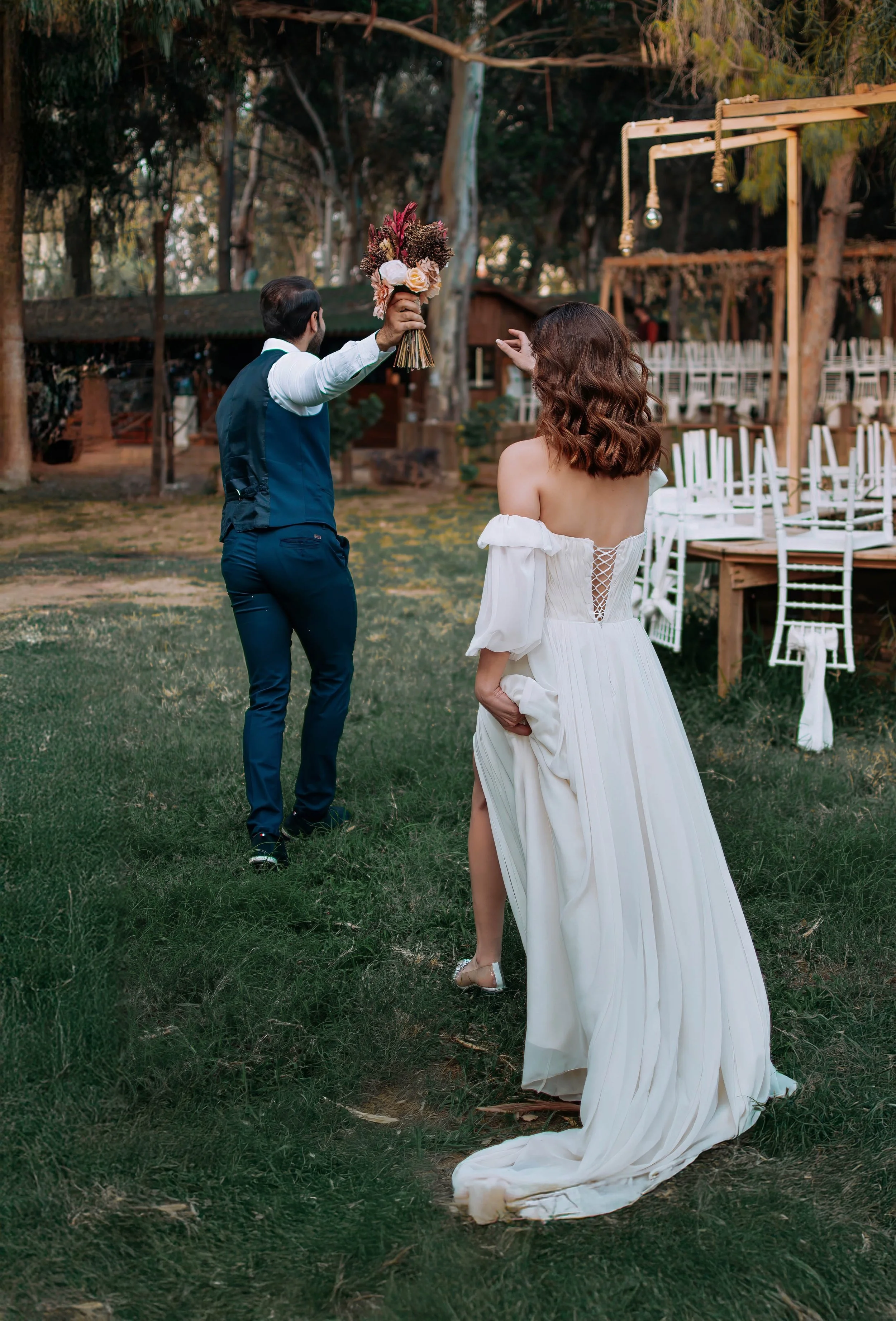 A woman in a white wedding dress stands outdoors on grass, holding up her dress, with a man in a vest and tie approaching her holding a bouquet of flowers. In the background, there are chairs and a wooden structure, and tall trees surrounding the are