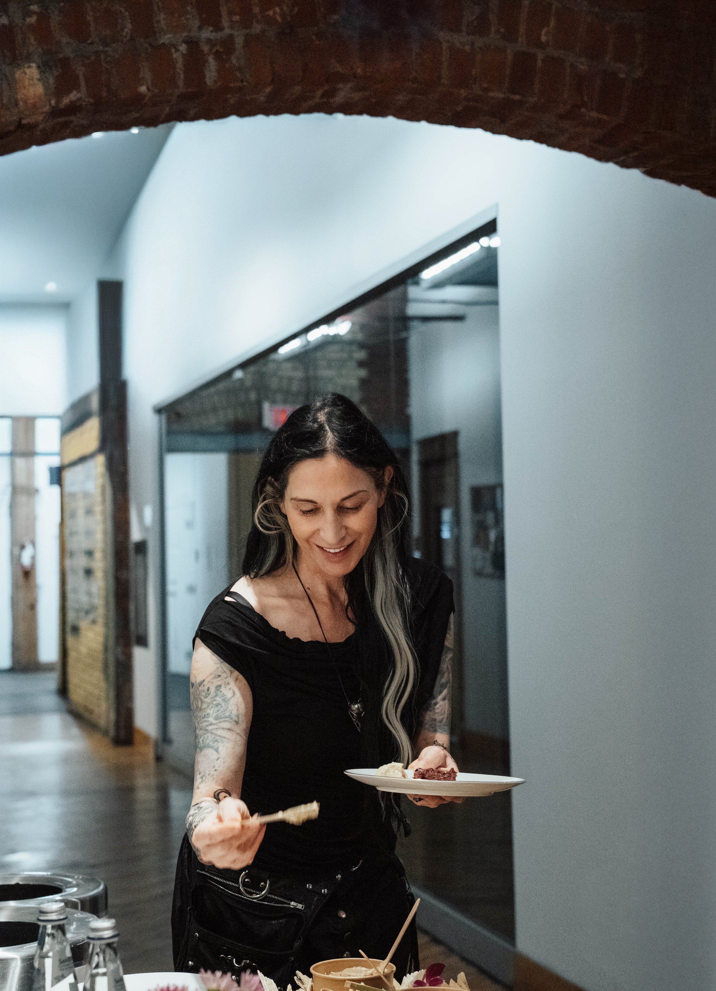 Woman with tattoos, dressed in black, smiling and serving herself food at an indoor gathering.