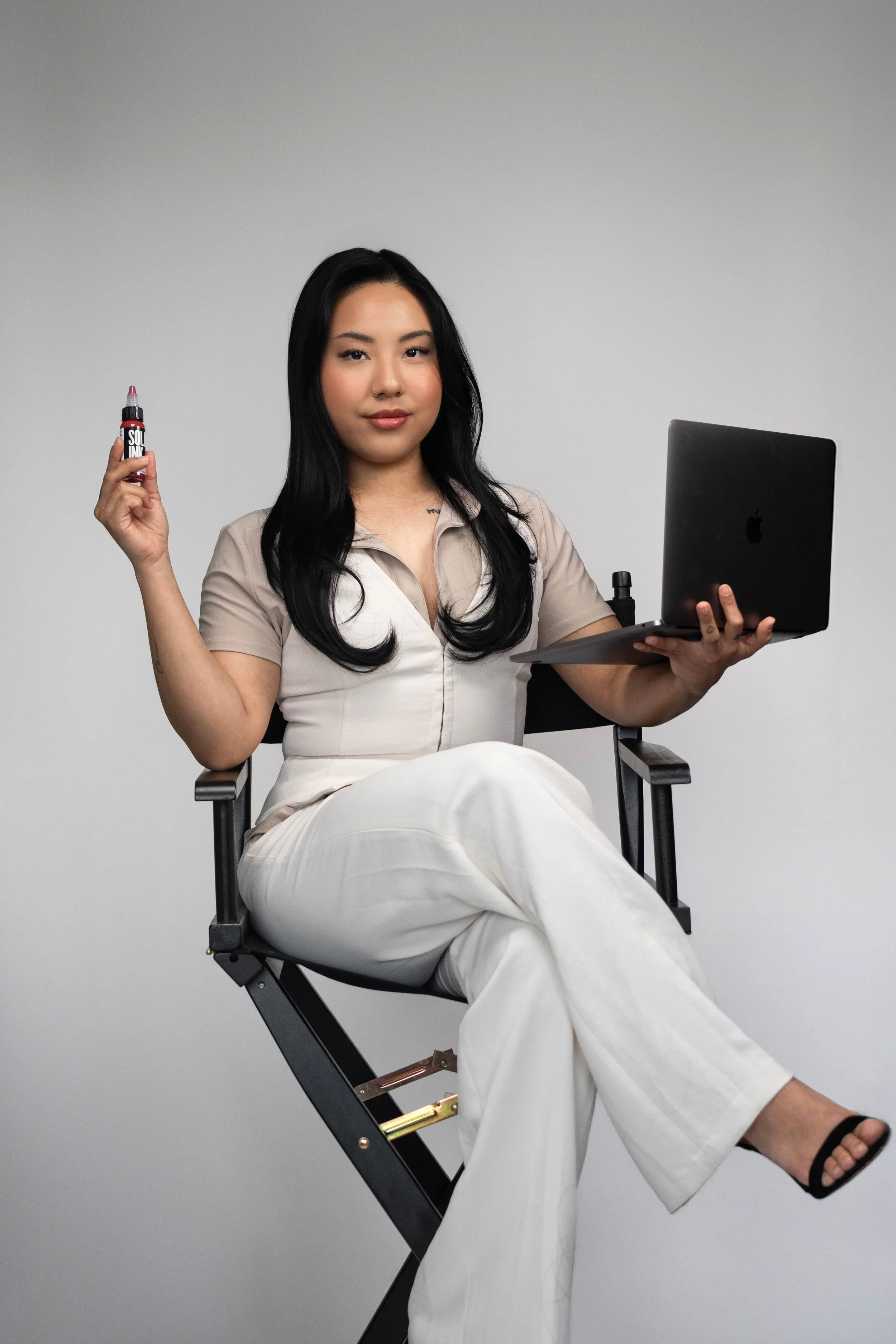 A woman sitting on a director's chair holding a makeup product in one hand and a laptop in the other, against a plain background.