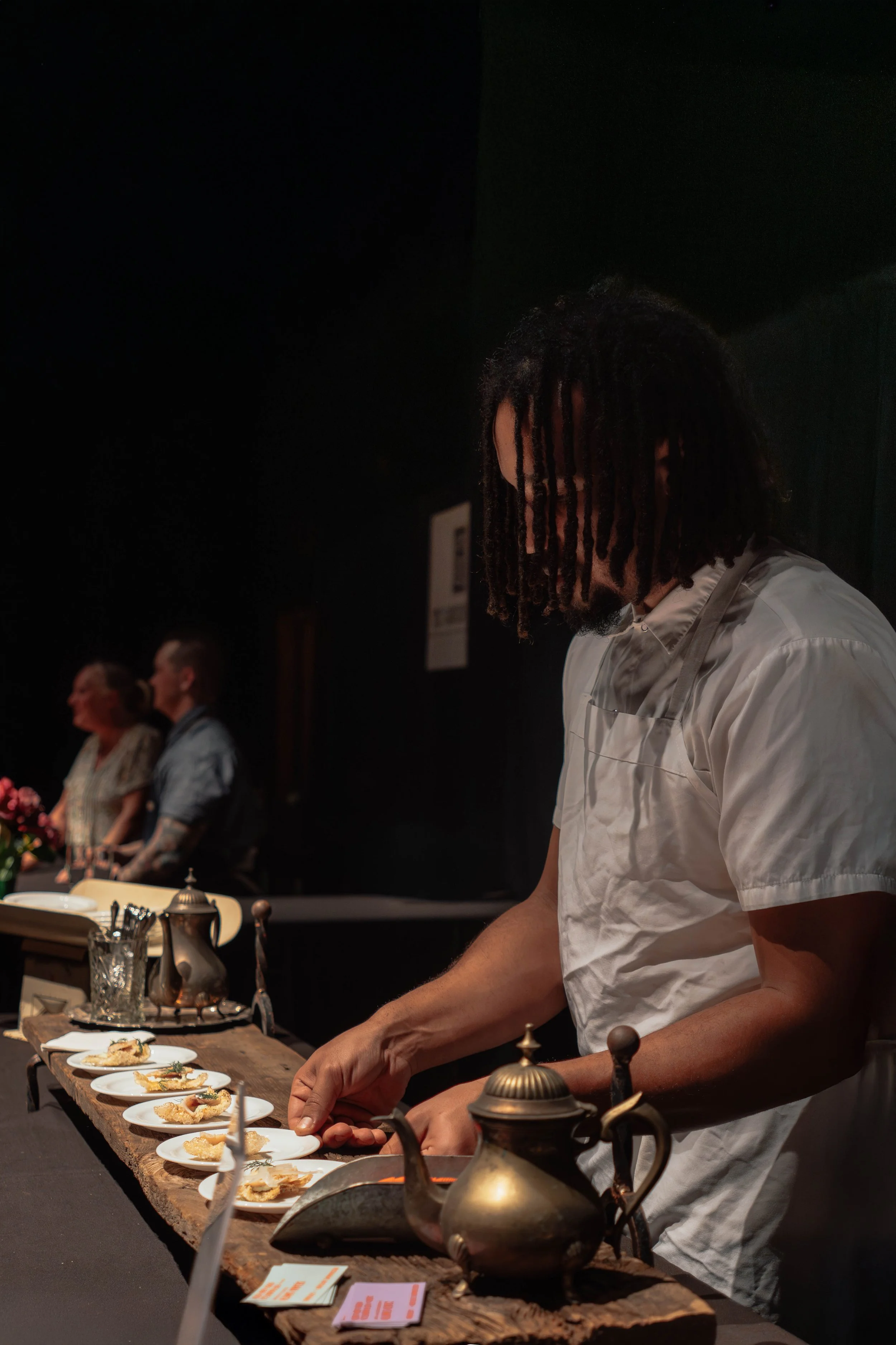 A chef with dreadlocks, wearing a white shirt and apron, standing at a wooden table with small plates of appetizers, a teapot, and decorative items, preparing food in a dimly lit setting.