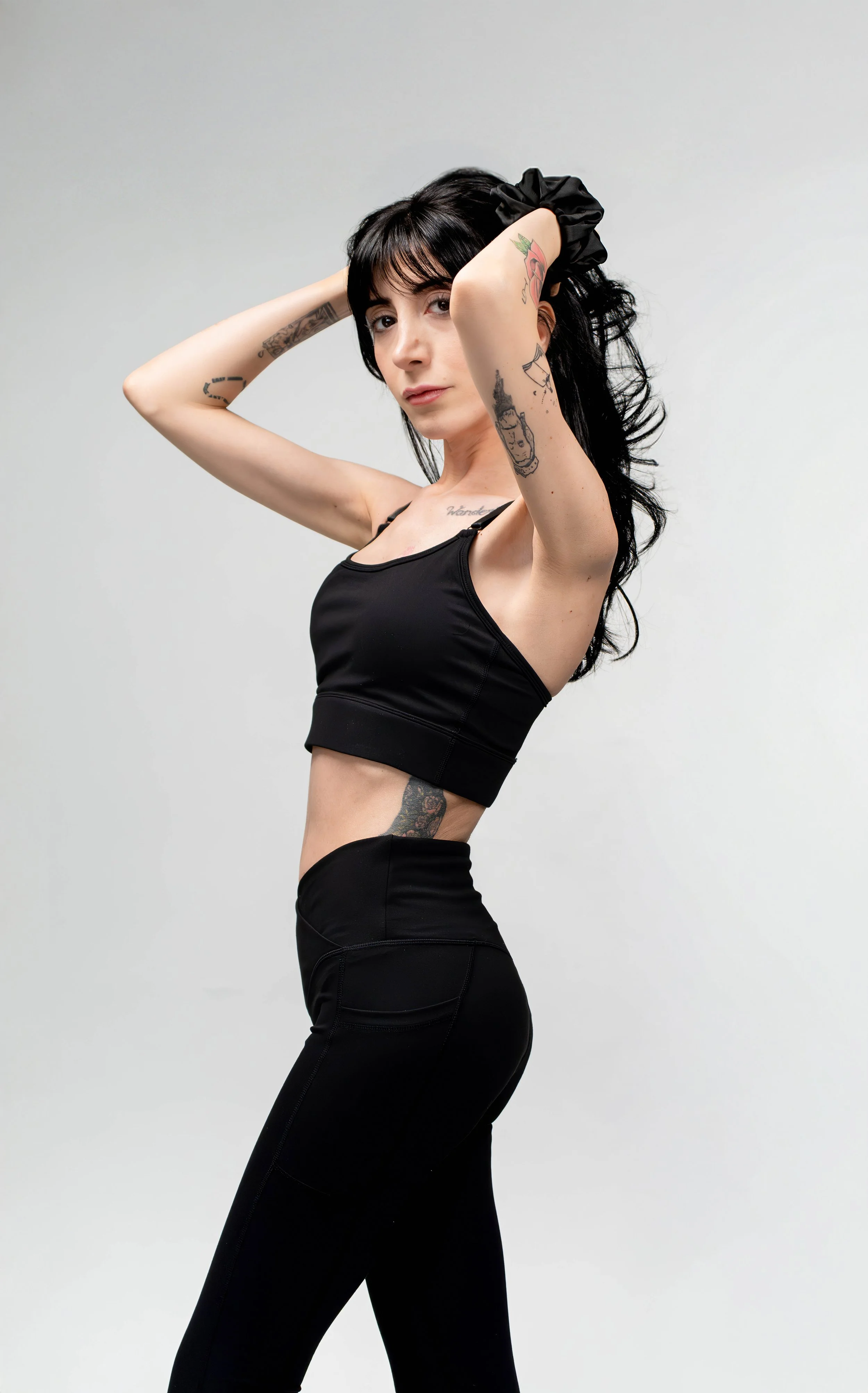 A young woman with black hair and tattoos wearing a black sports bra and black leggings poses against a plain white background.