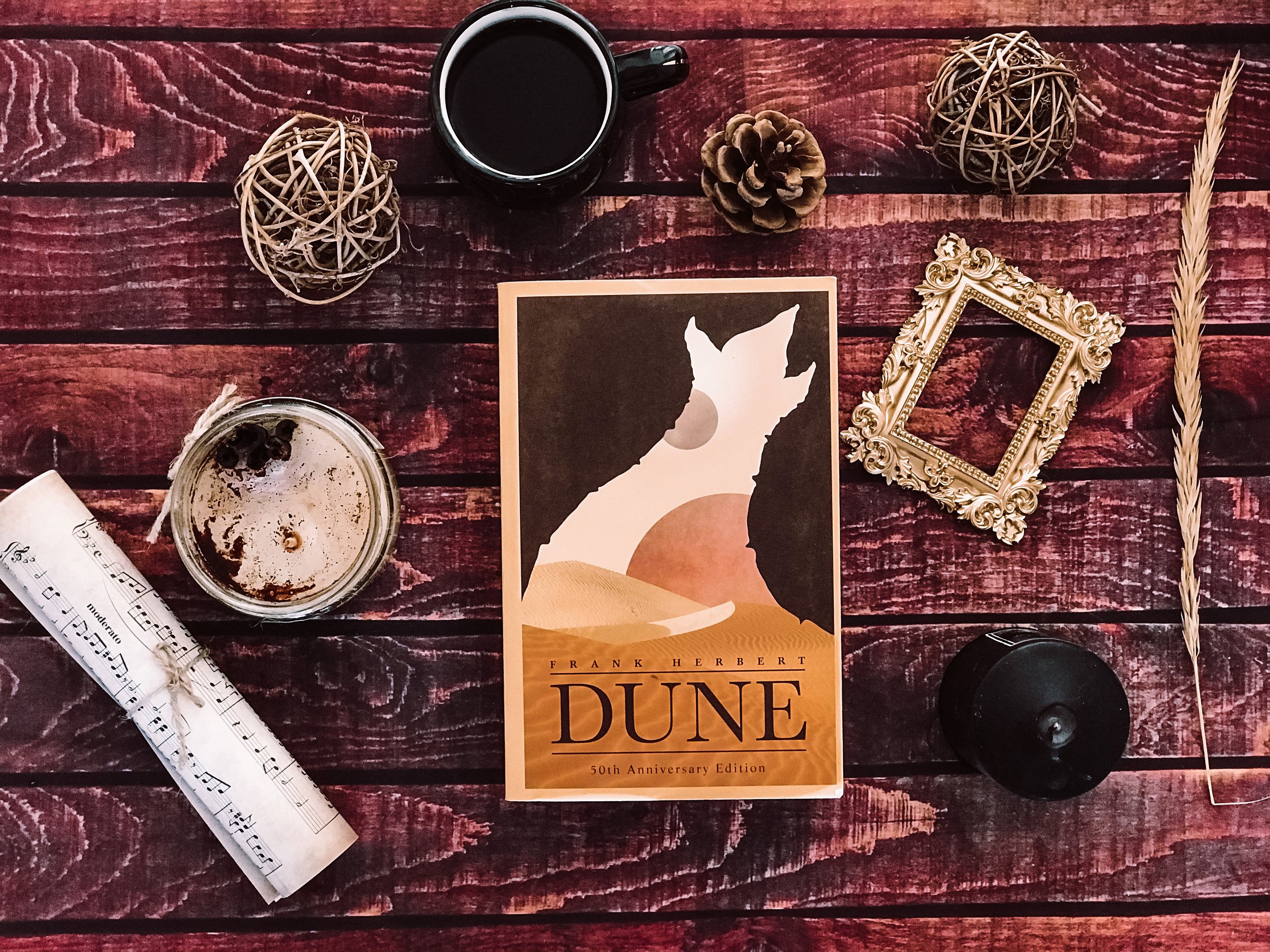 A flat lay of a wooden table with a mug of black coffee, a glass jar with a drink and berries, a rolled sheet of music, a book titled Dune, decorative wicker balls, an ornate picture frame, a pine cone, and a dried wheat stalk.