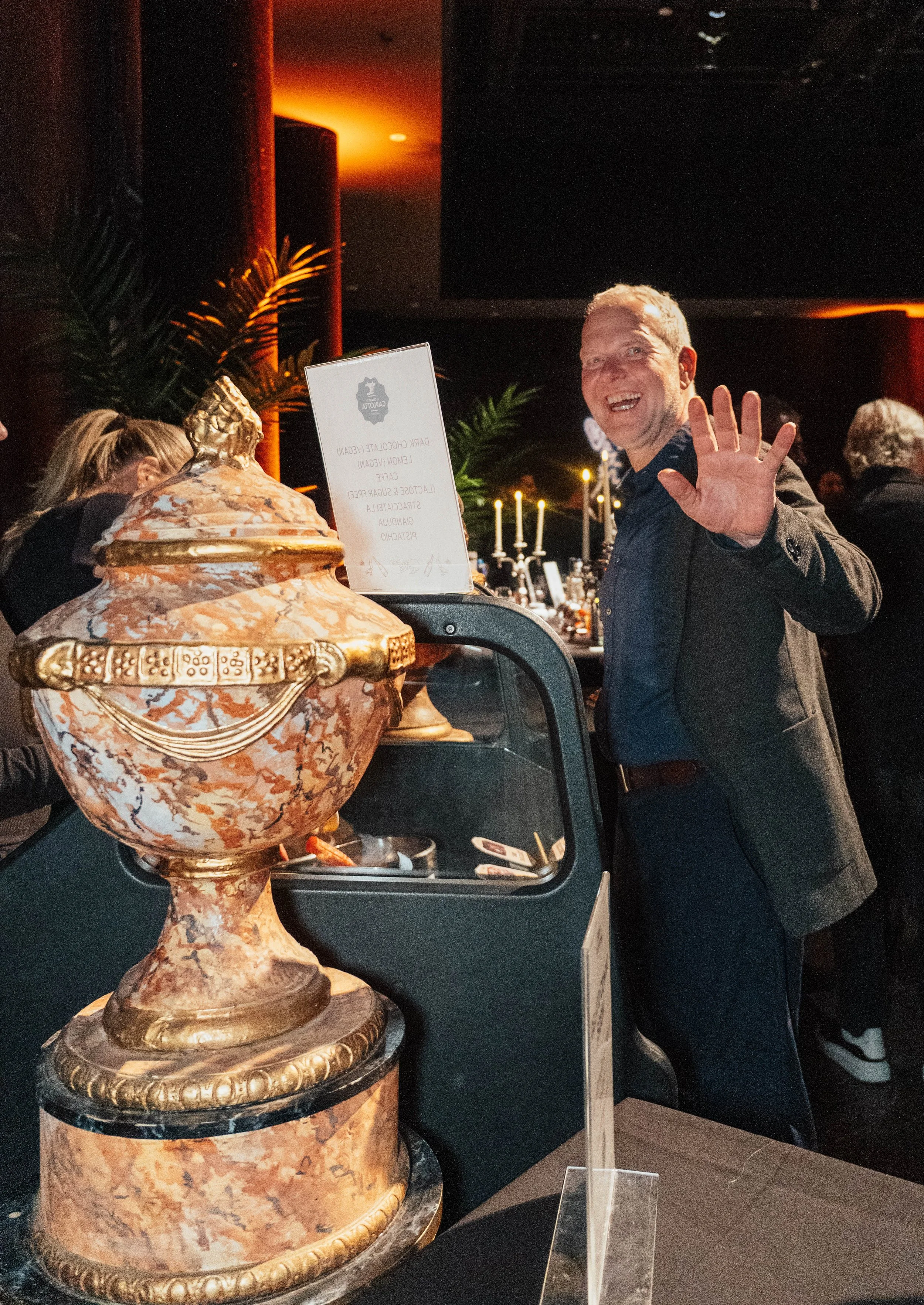 A man waves and smiles at a buffet table with a large ornate ceramic urn in the foreground, in a dimly lit banquet hall with candlelit decor.