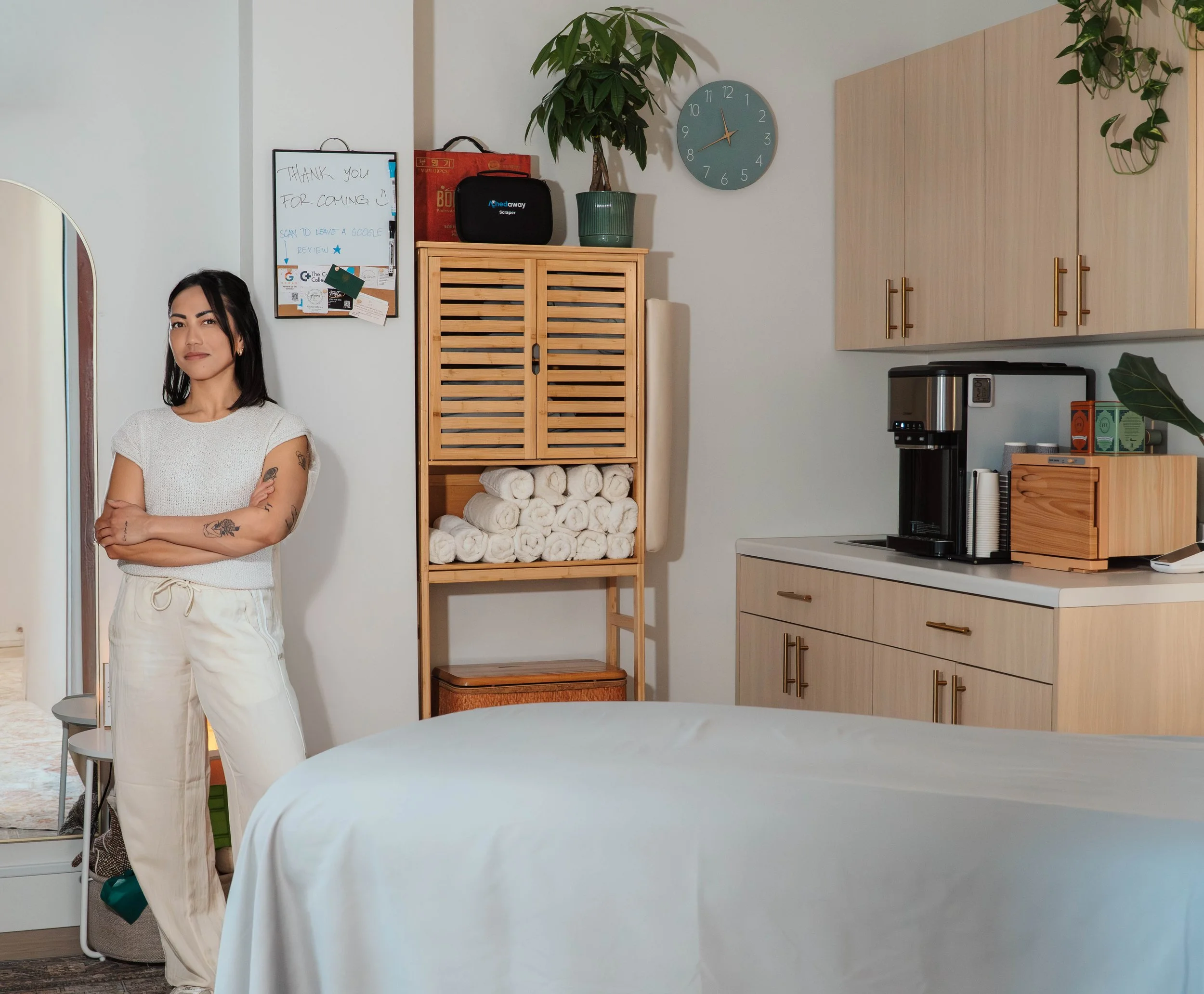 A woman with tattoos on her arms standing with arms crossed near a bed in a modern, neatly organized room with wooden cabinets, a wall clock, a whiteboard with notes, and a coffee maker on a cabinet.