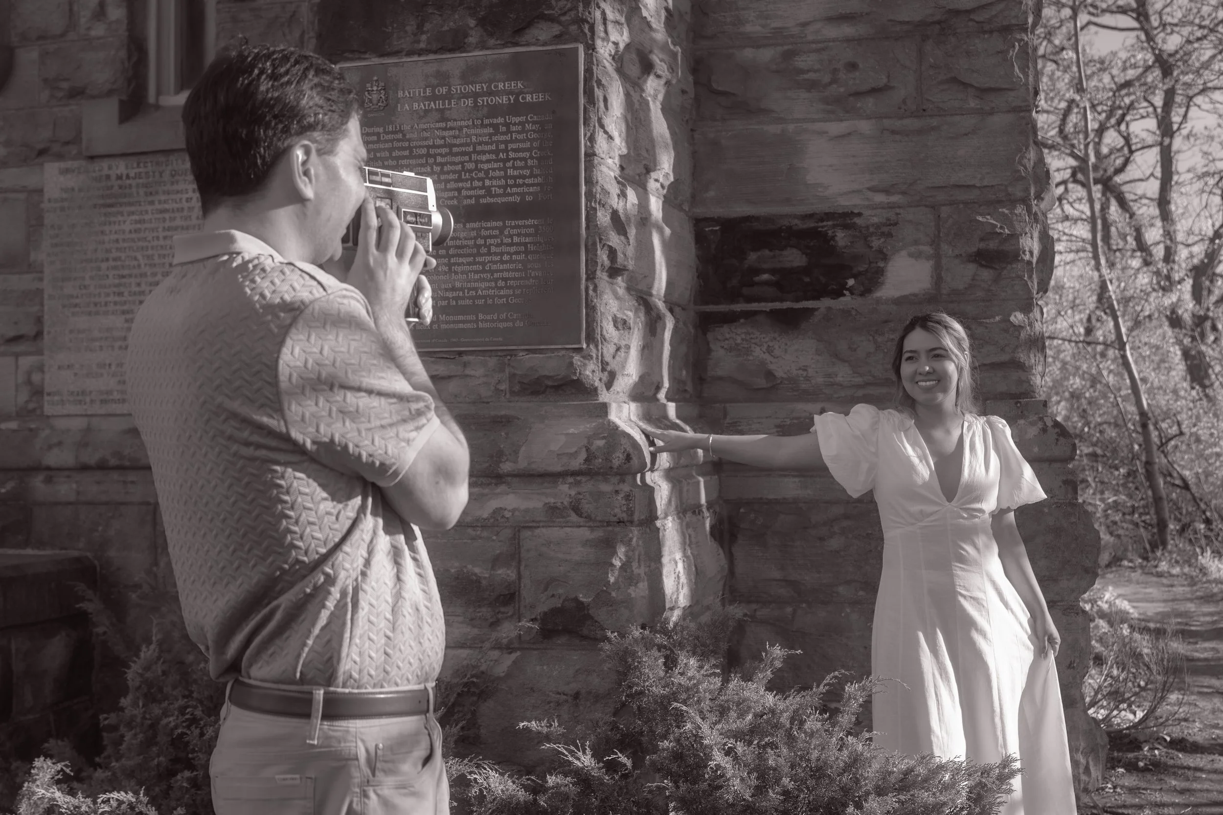 A man taking a photograph of a woman leaning against a stone wall outside with trees in the background.