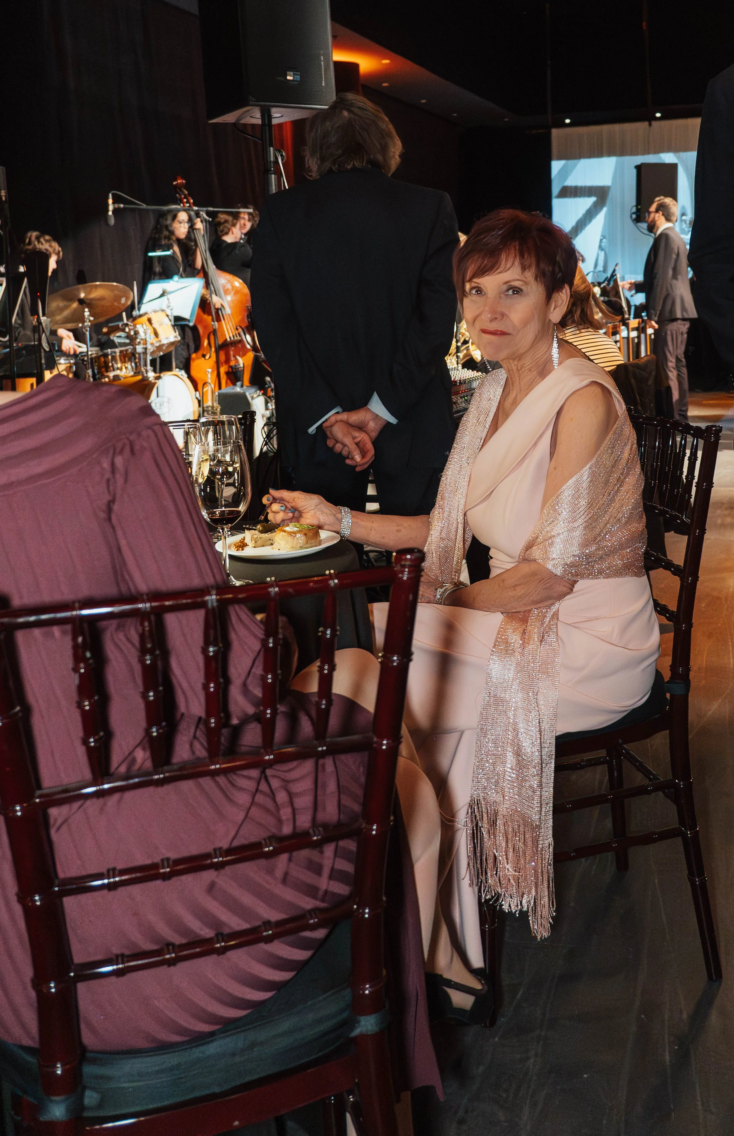 A woman in a light pink dress and sparkling shawl sits at a formal event, eating dessert. She is at a table with wine glasses, and there are musicians playing in the background.