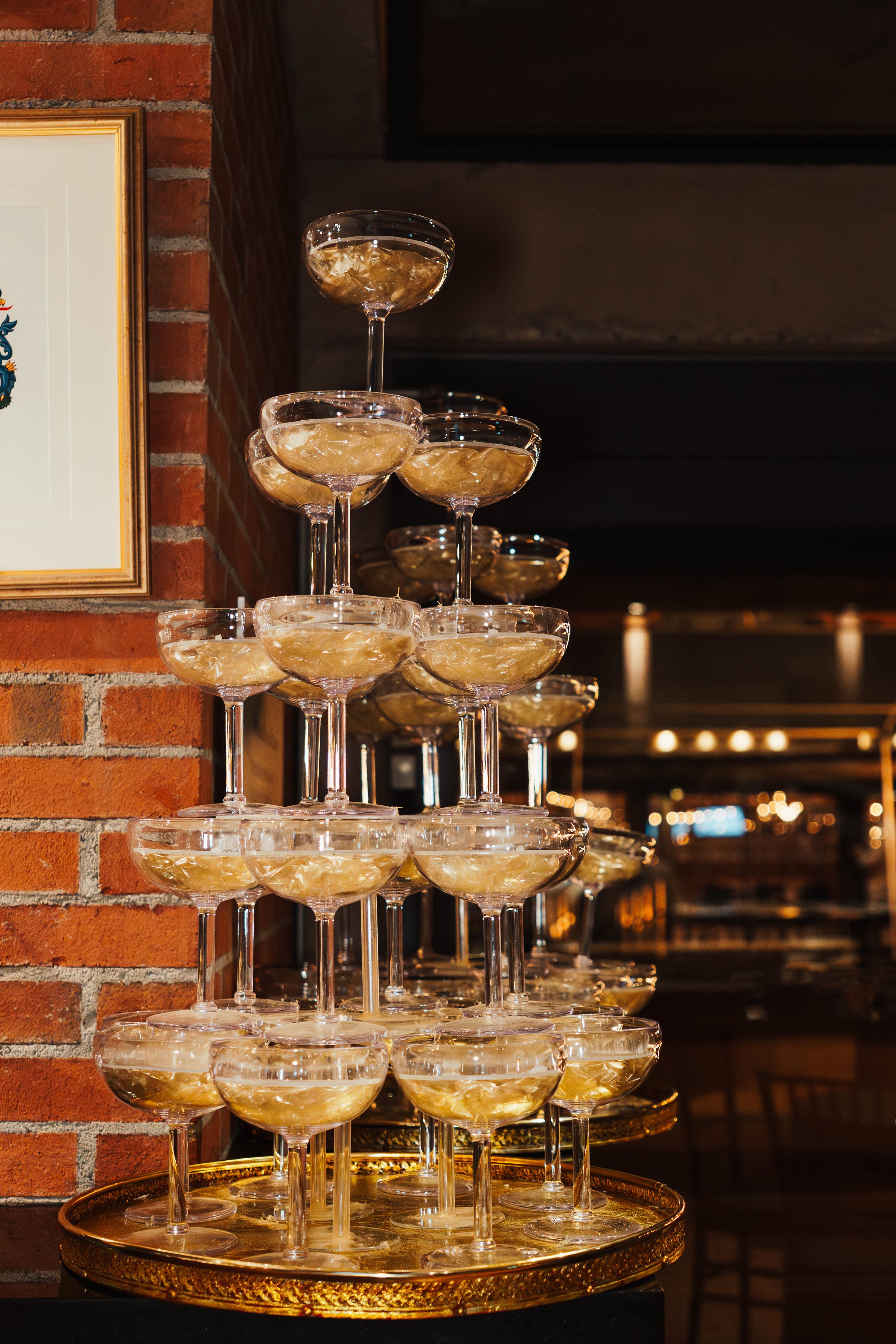 A pyramid of empty cocktail glasses with gold rims, stacked on a gold tray, against a brick wall in a dimly lit bar or restaurant.