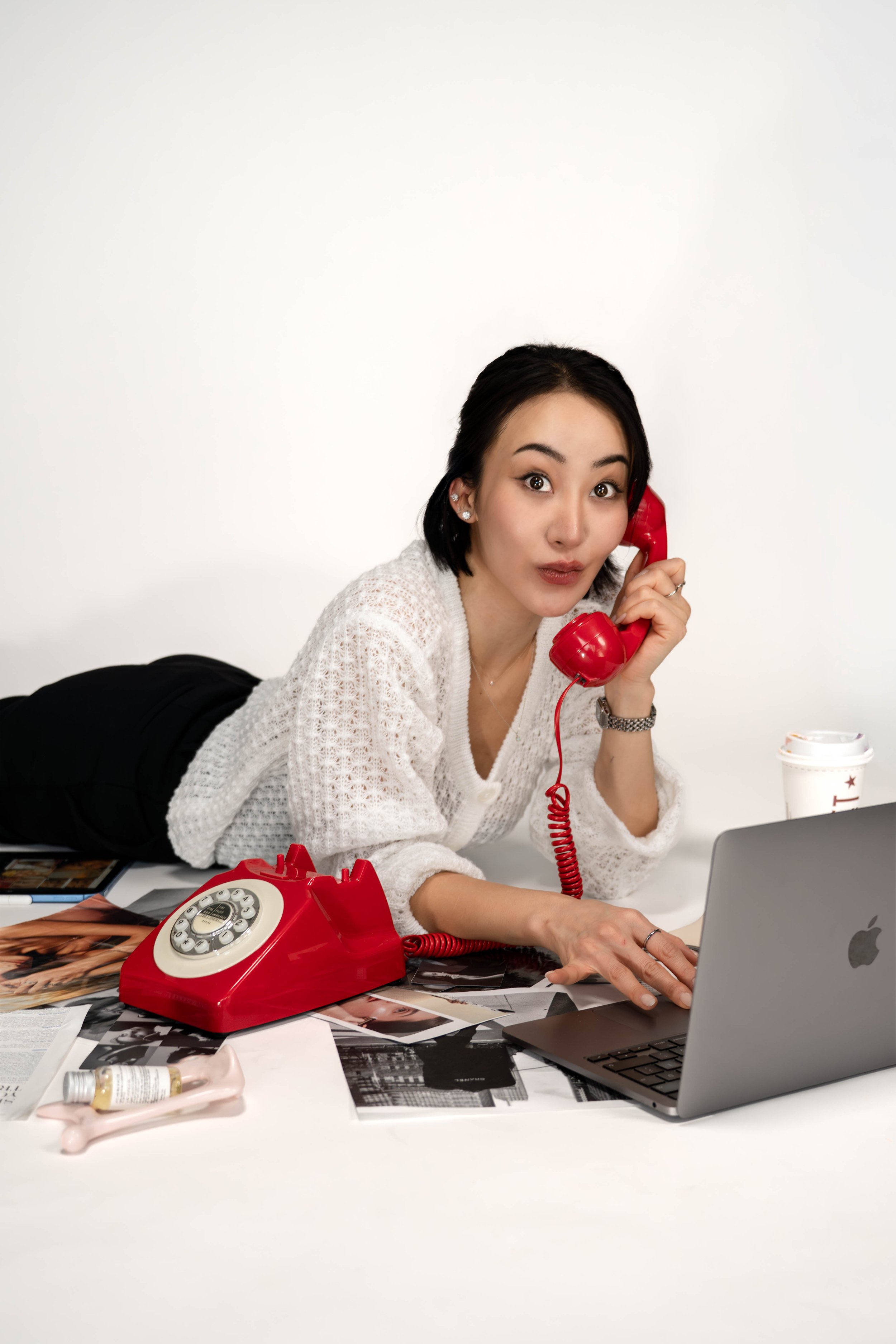 A woman lying on her stomach on a desk, talking on a red vintage rotary phone, with a laptop and scattered magazines on the desk.