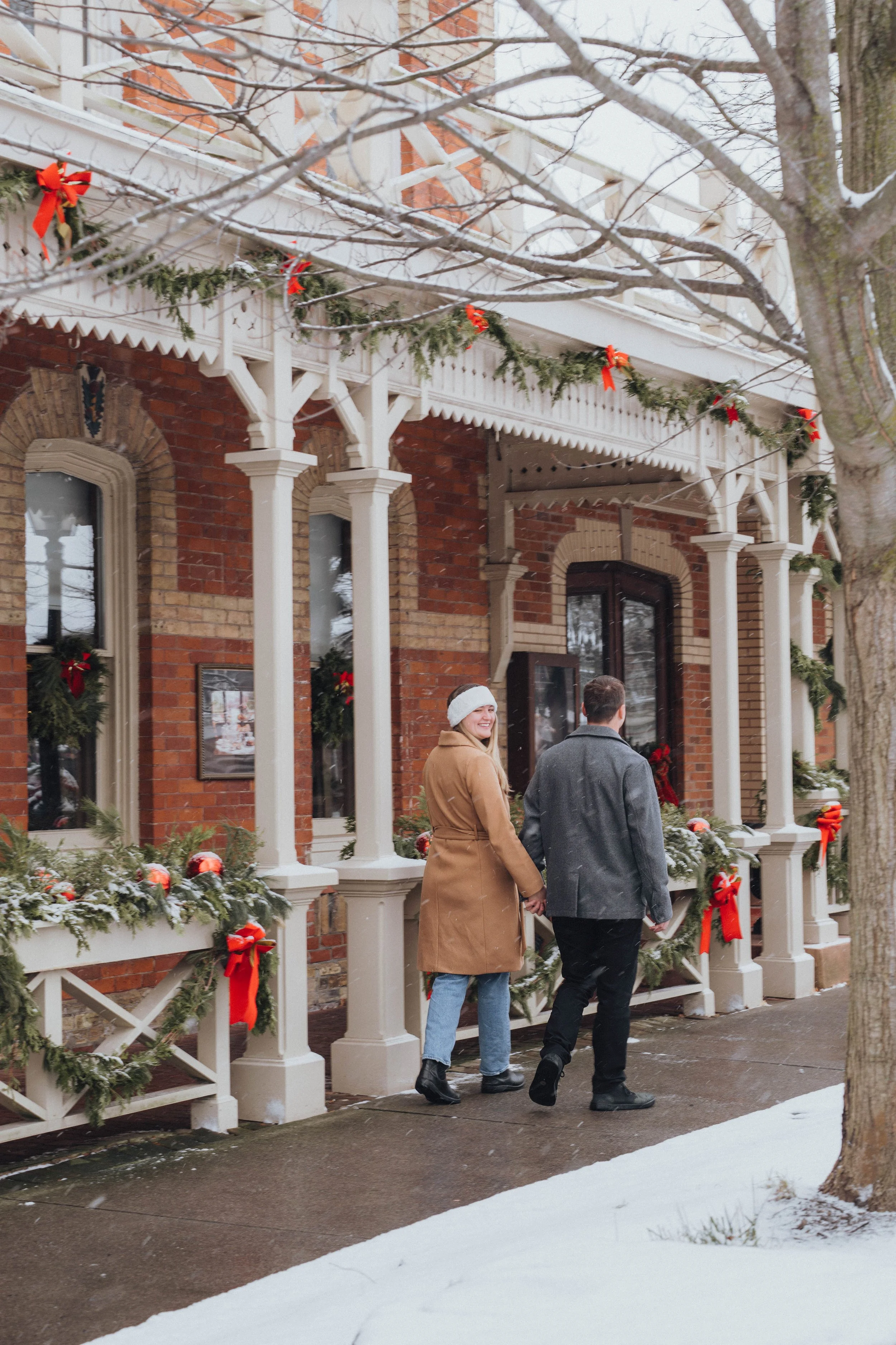 Christmas couple photography in Niagara-on-the-Lake, walking hand in hand past historic homes decorated with festive greenery and red bows.