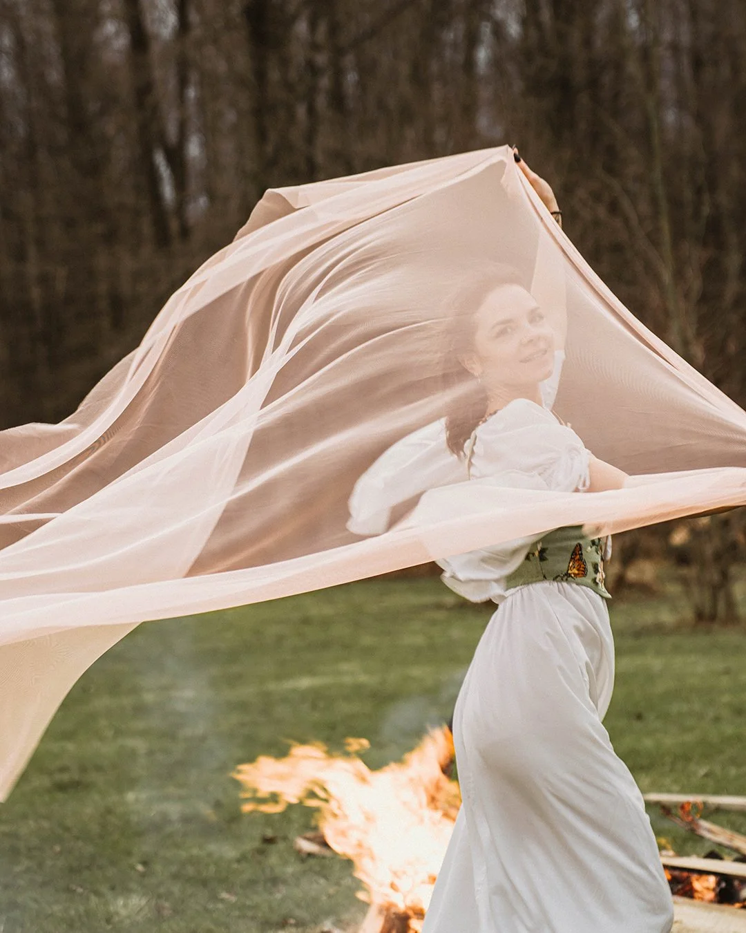 A woman dressed in a white dress with a butterfly belt stands outdoors on grass, with a semi-transparent fabric draped around her, and a small fire burning on the ground behind her.