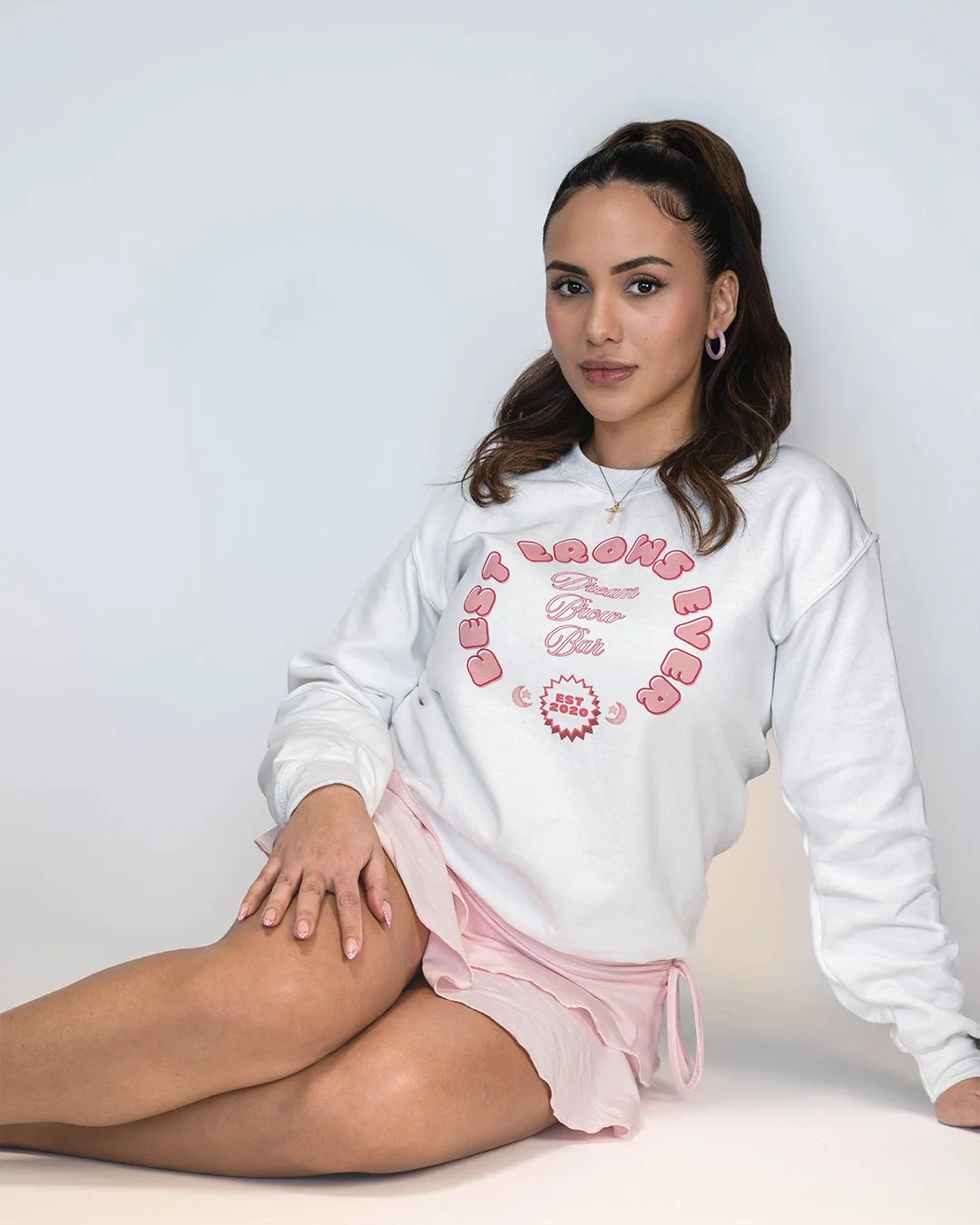 Young woman with brown hair in a high ponytail, wearing a white sweatshirt with pink text and graphics, pink shorts, and purple hoop earrings, sitting on the floor against a plain white background.