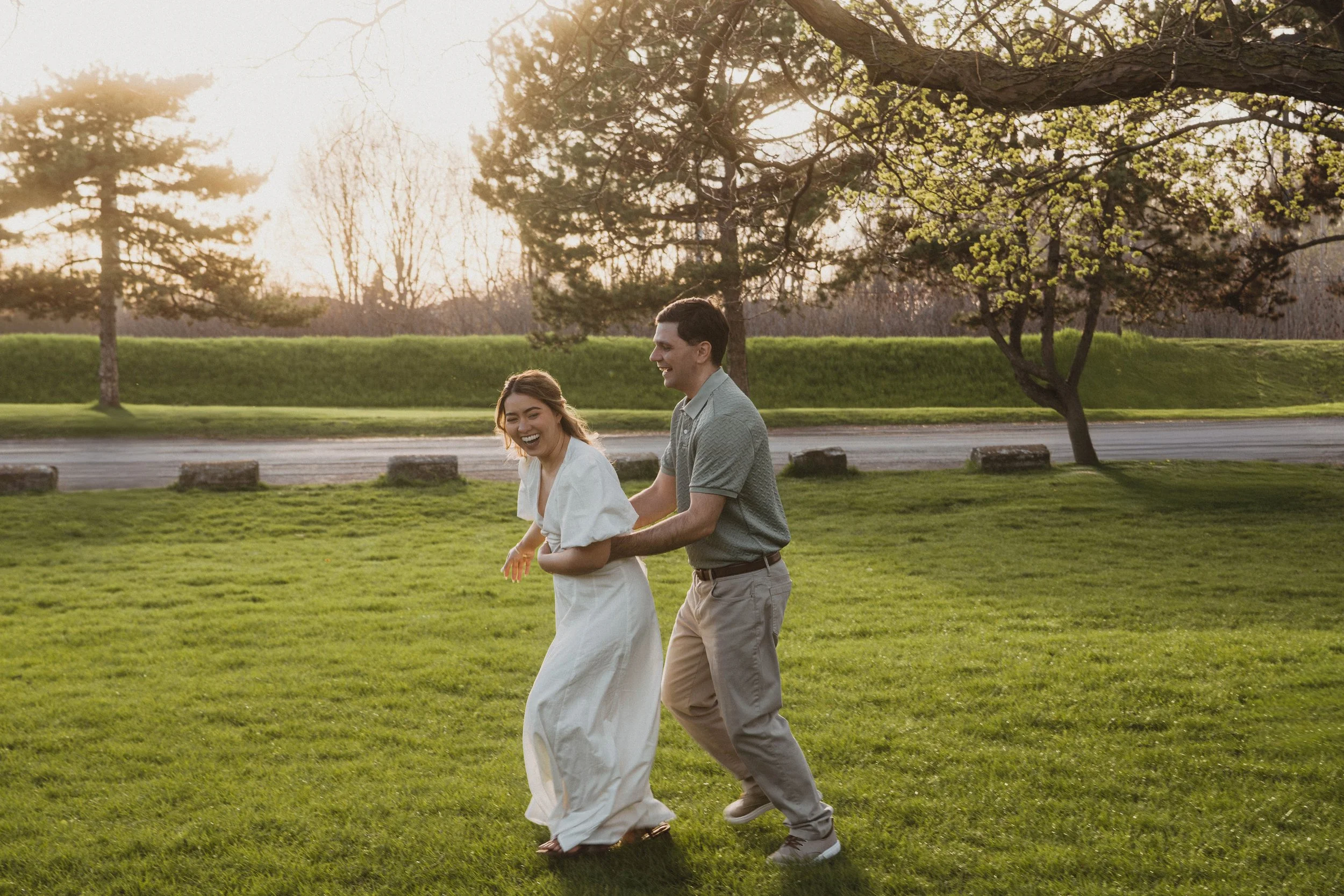 A couple is joyfully holding hands and laughing while walking together outdoors in a park-like setting during sunset, surrounded by green grass, trees, and a road in the background.