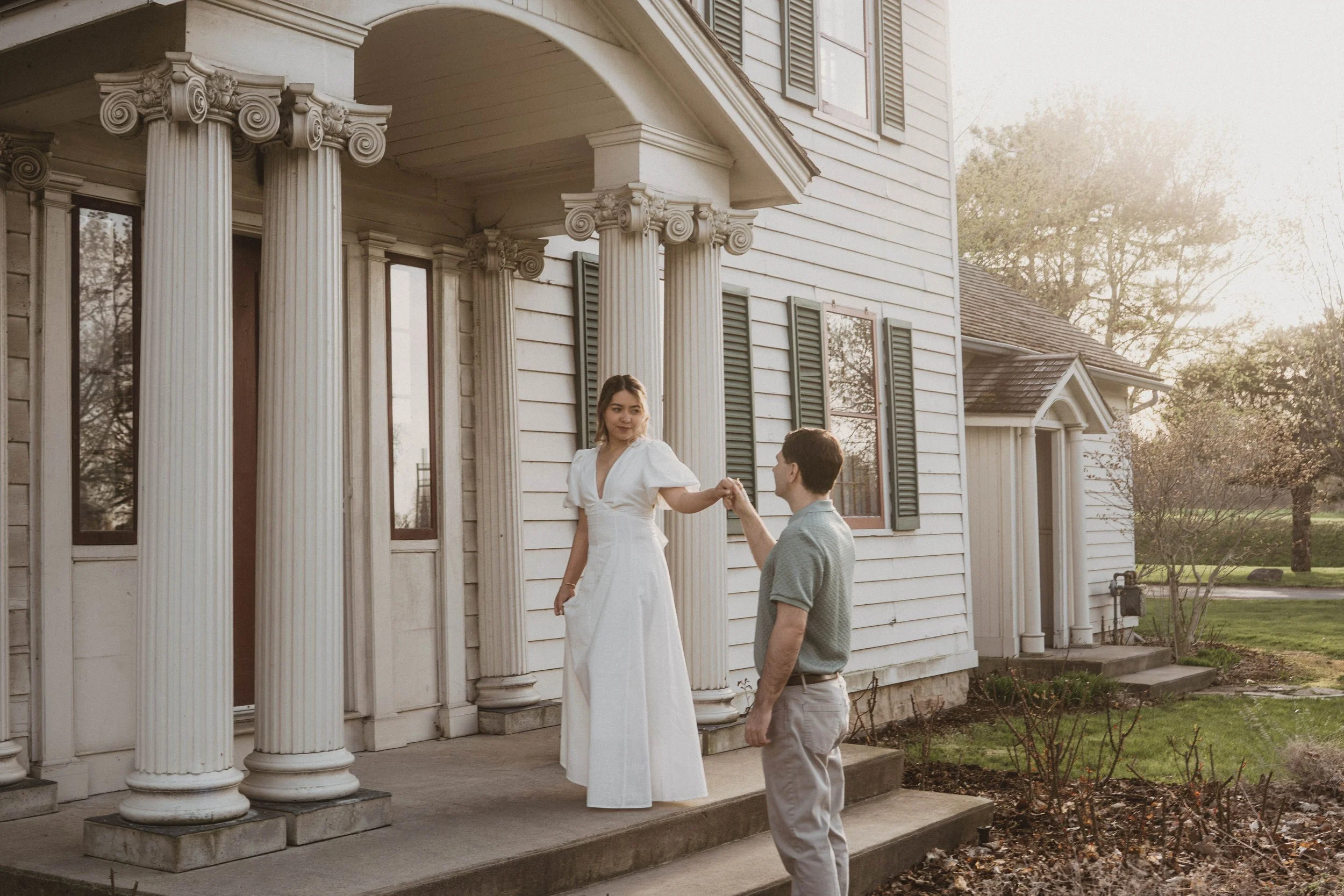 A woman in a white dress kneeling on one knee, holding hands with a man in a gray polo shirt, outside in front of a white house with classical columns, during sunset.