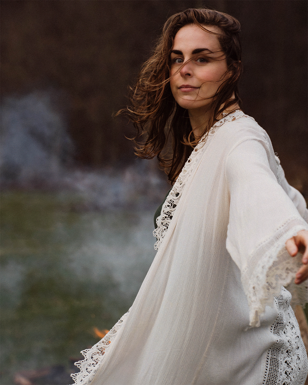 A woman with long brown hair standing outdoors near a body of water with steam rising from it, wearing a light-colored, lace-trimmed garment, looking at the camera.