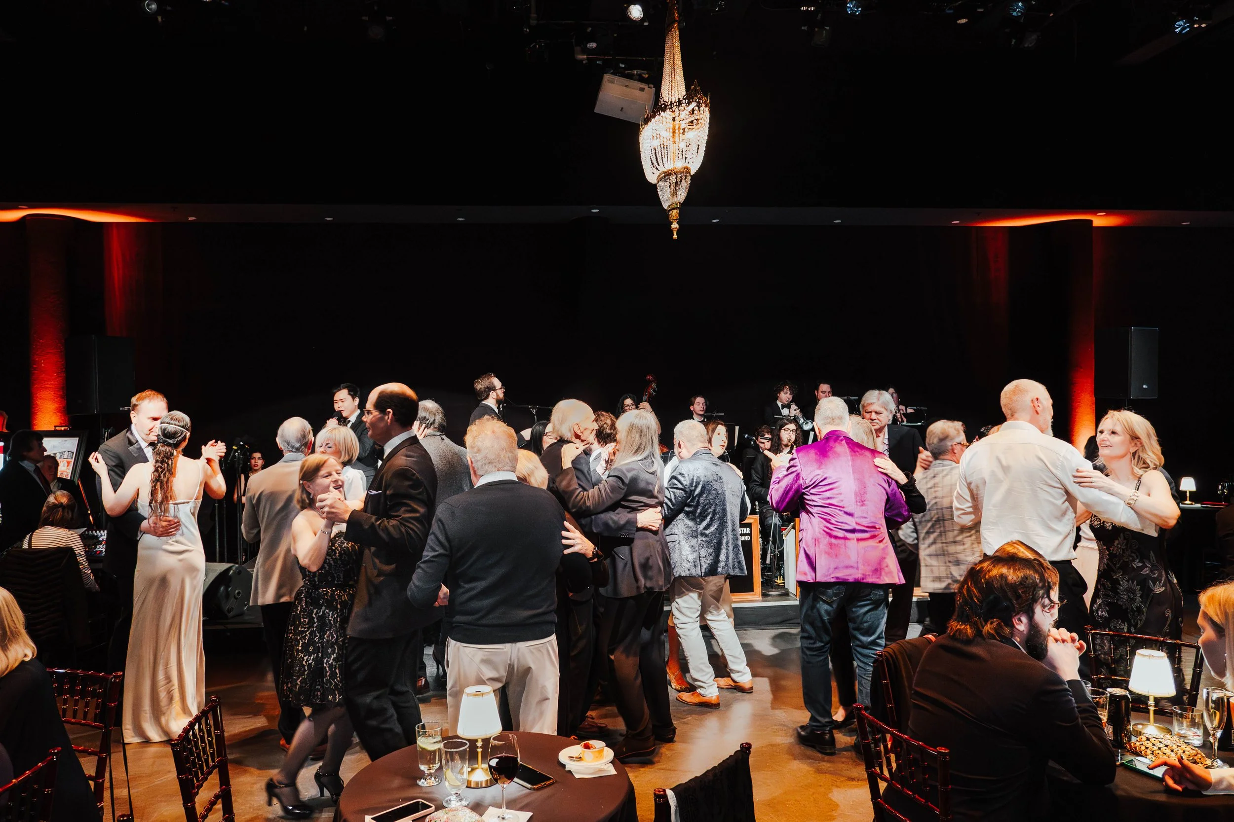 People dancing and socializing at an evening jazz event in a dimly lit venue with a chandelier, musicians playing on stage, and tables with drinks and appetizers.