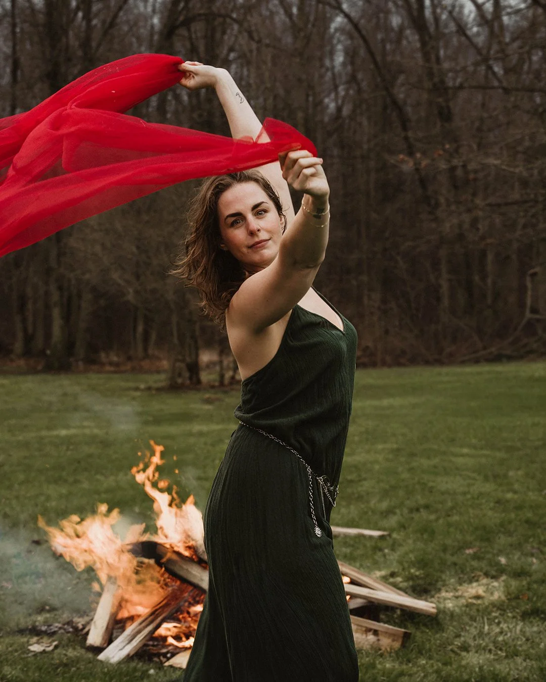 A woman with shoulder-length brown hair waving a red cloth in the air outdoors near a fire on a wooden structure in a grassy field with leafless trees in the background.