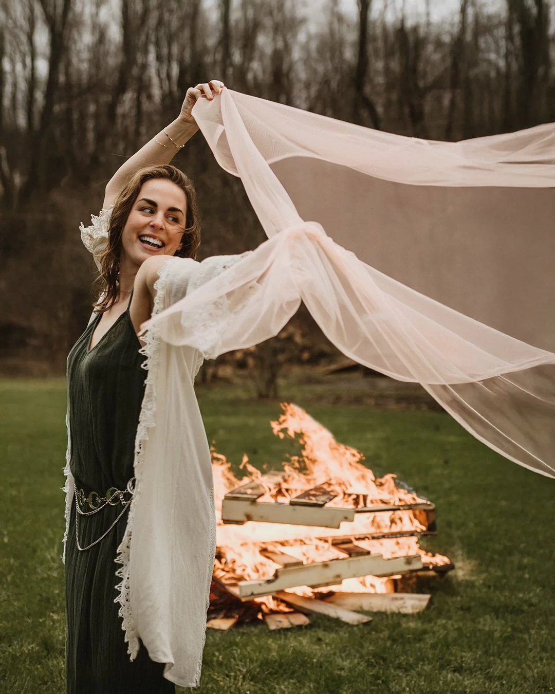A woman smiling, holding a light pink cloth or veil, standing outdoors on grass during dusk. Behind her, a fire is burning on a wooden pallet.