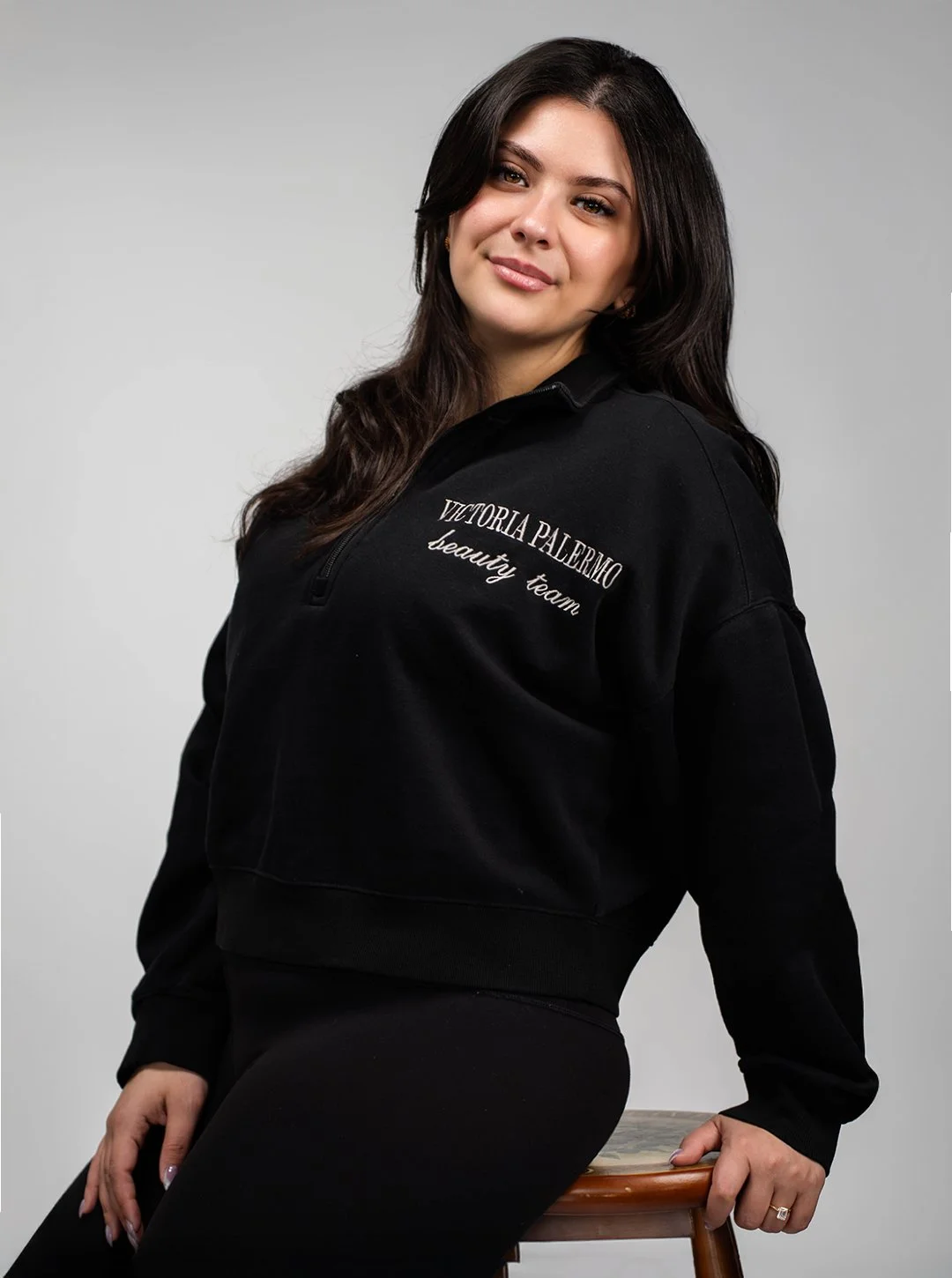 A woman with dark brown hair and light skin wearing a black zip-up sweatshirt with the words "Victoria Palazzo beauty team" embroidered on it, sitting on a stool against a plain light gray background, looking at the camera and smiling slightly.