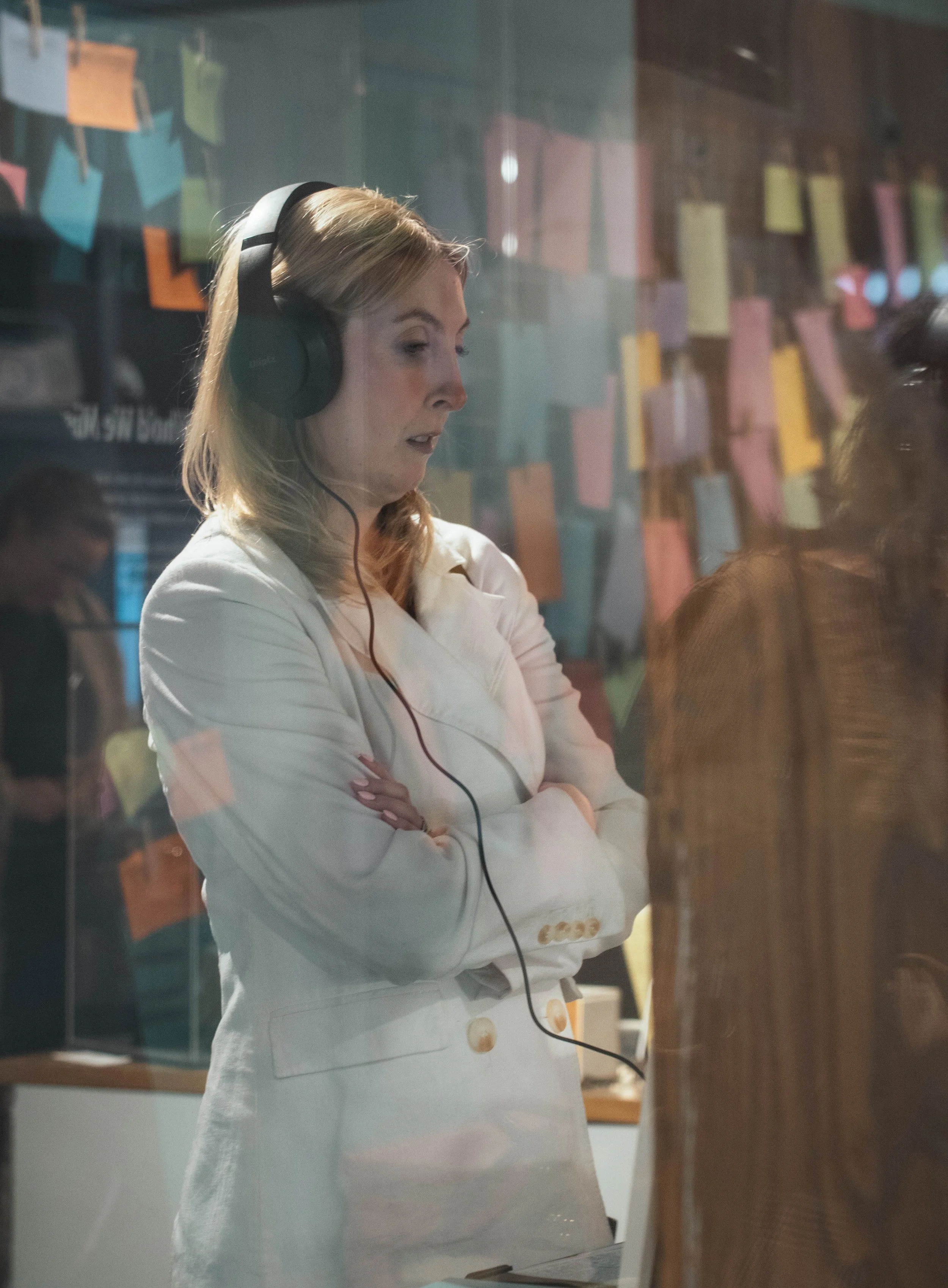 A woman wearing a white blazer with arms crossed, listening with headphones through a glass window in an office or studio with colored sticky notes on a wall in the background.