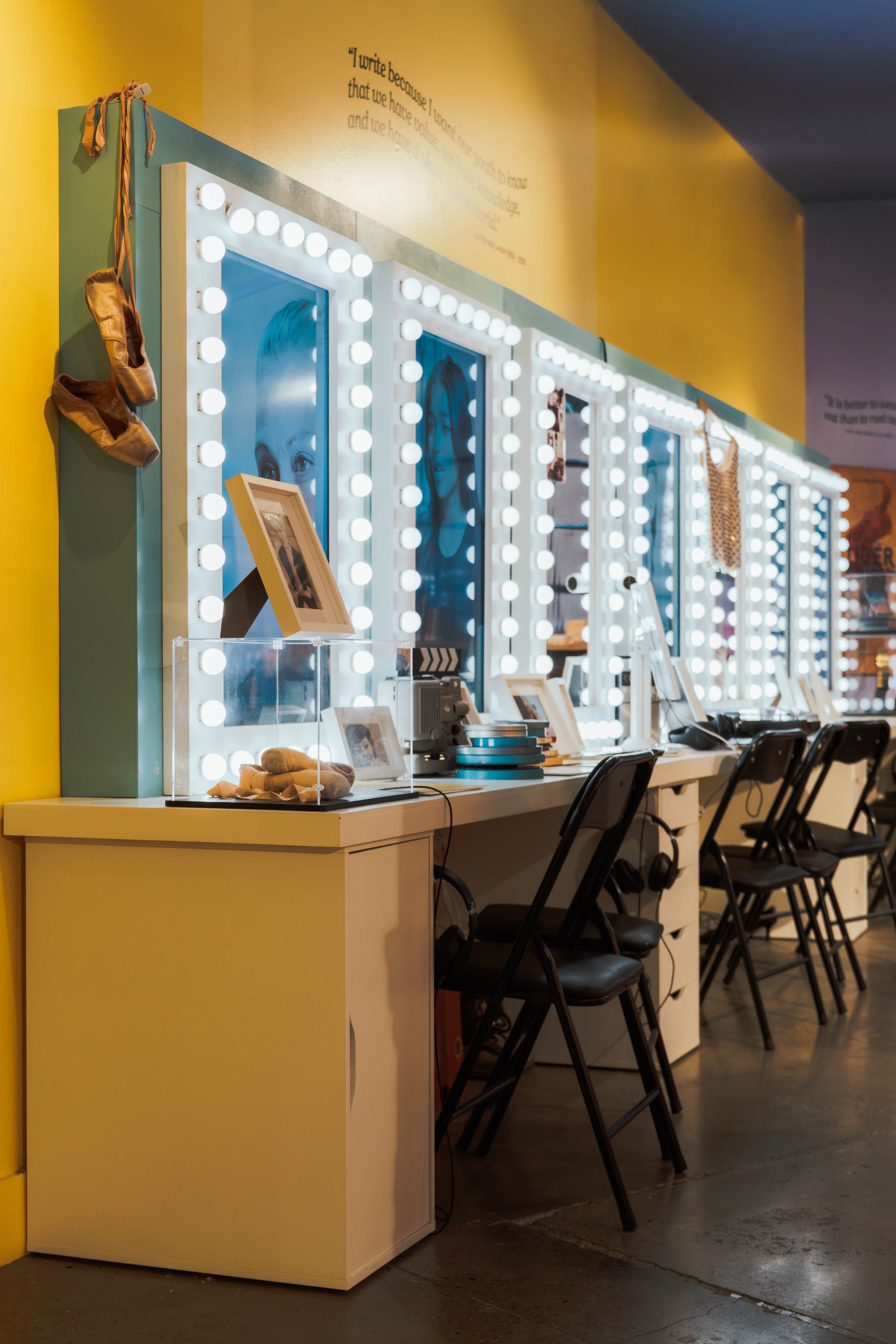 A brightly lit dressing room area with illuminated mirrors, framed photographs, ballet shoes hanging on the corner, and a counter with chairs.