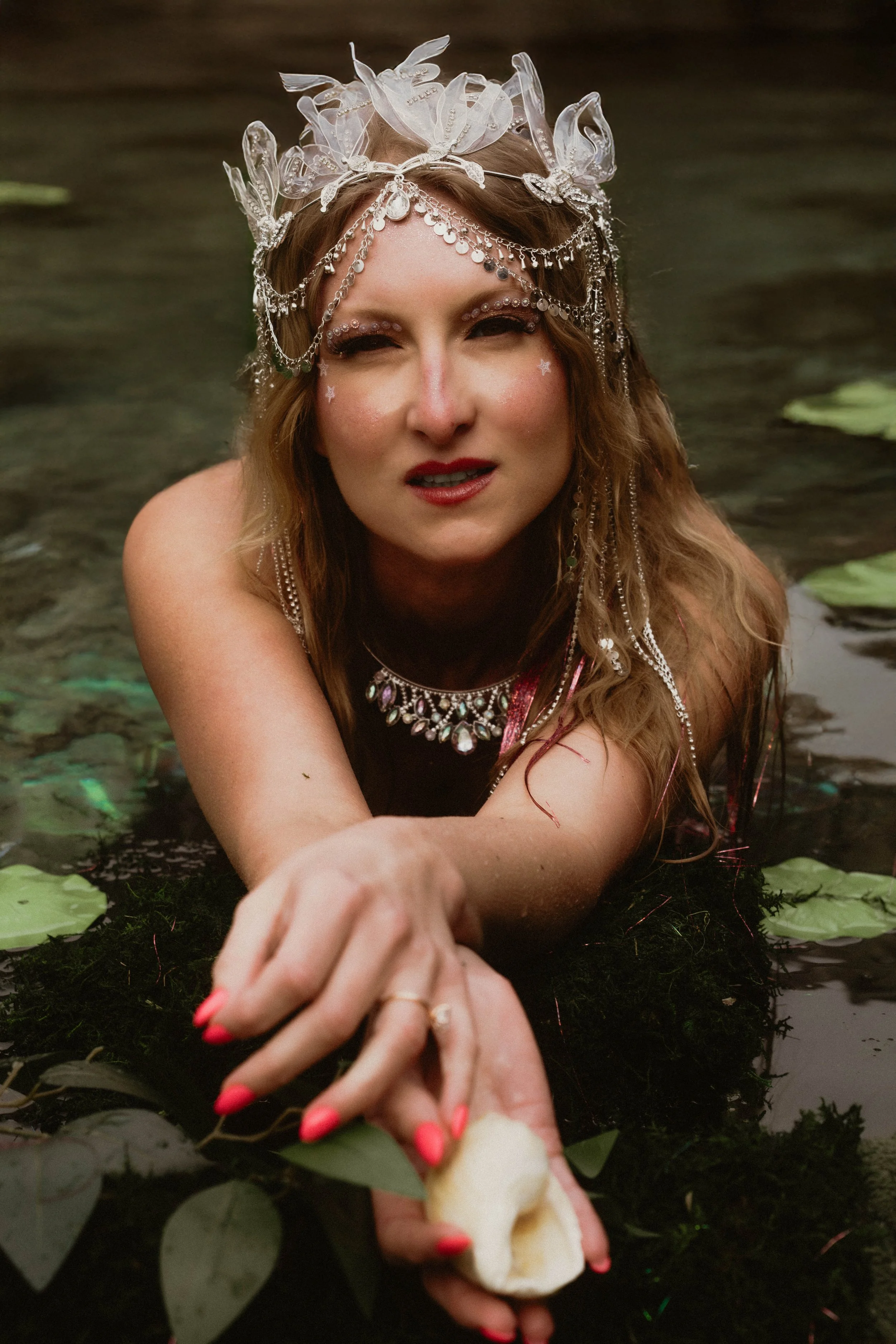 A woman with elaborate jewelry and a crown made of transparent plastic leaves and chains, lying in water surrounded by lily pads, reaching out her hand with painted red nails.